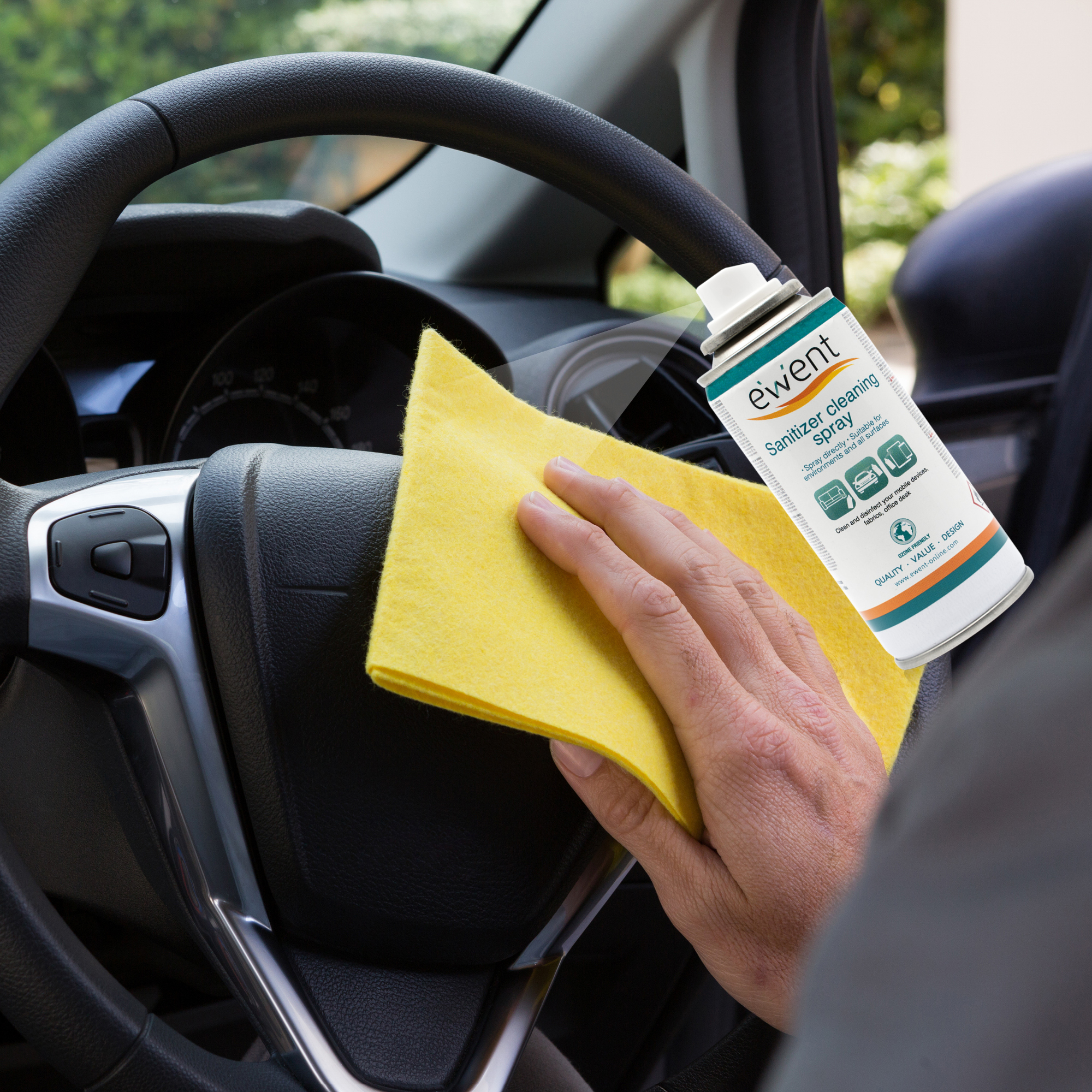 Ręka czyści kierownicę samochodu żółtą szmatką. W pobliżu znajduje się spray dezynfekujący.