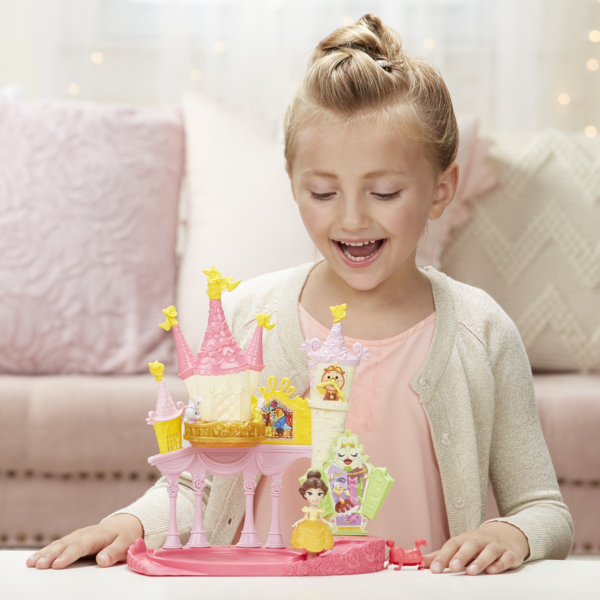 Una bambina sorride a un castello giocattolo. Il castello è rosa e giallo, con statuette di personaggi. Lo sfondo include un divano.