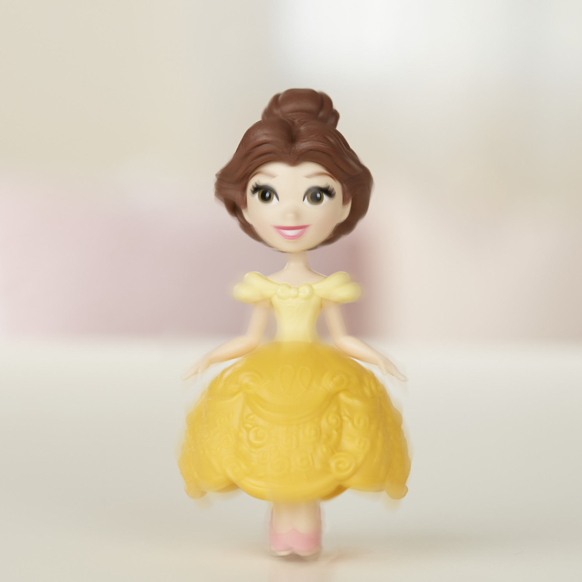 Una bambola di Belle indossa un vestito giallo. I suoi capelli castani sono raccolti. Lo sfondo è sfocato rosa e bianco.
