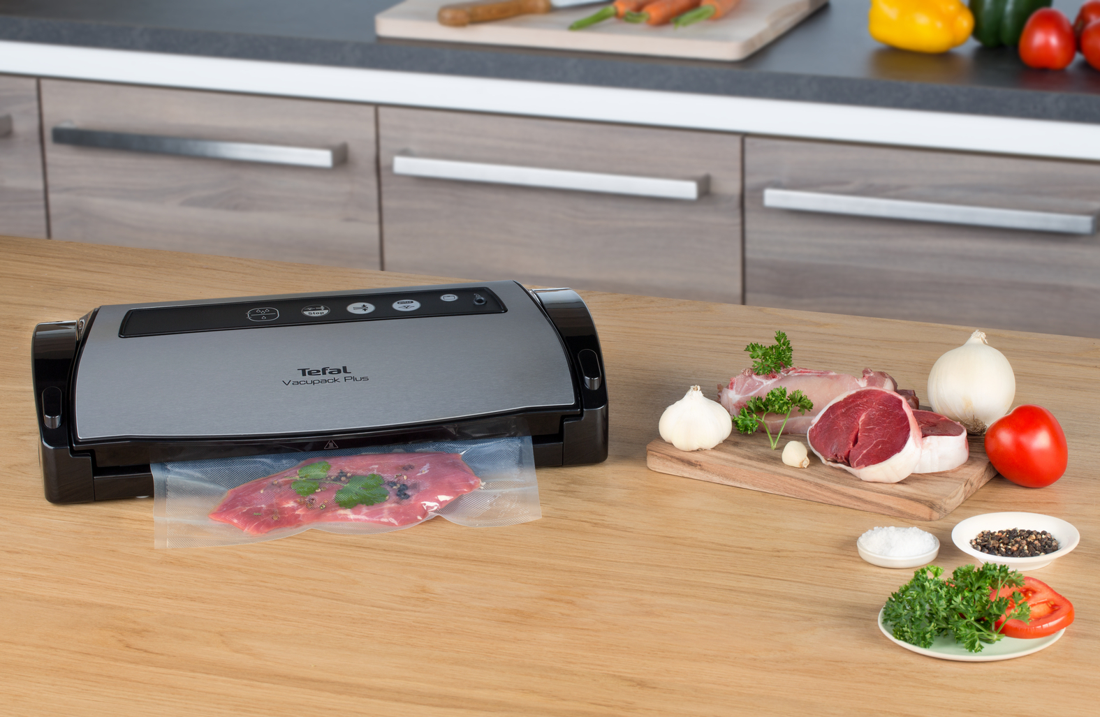 Zgrzewarka próżniowa z mięsem w worku, obok surowego mięsa, cebuli, pomidora, czosnku, pietruszki i przypraw, na drewnianym stole.