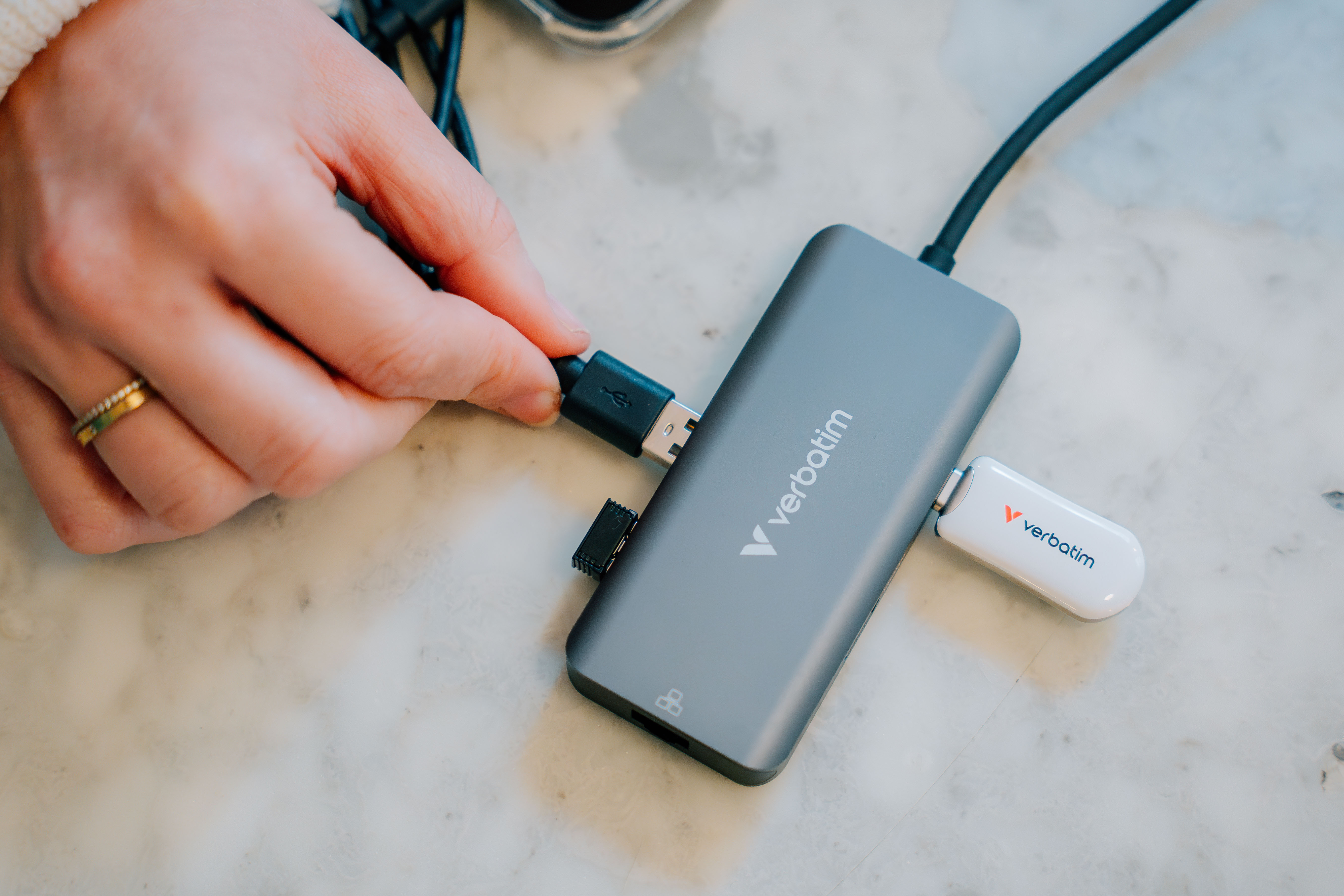 Hand plugging a USB cable into a grey Verbatim hub. A white Verbatim USB stick is also plugged in.