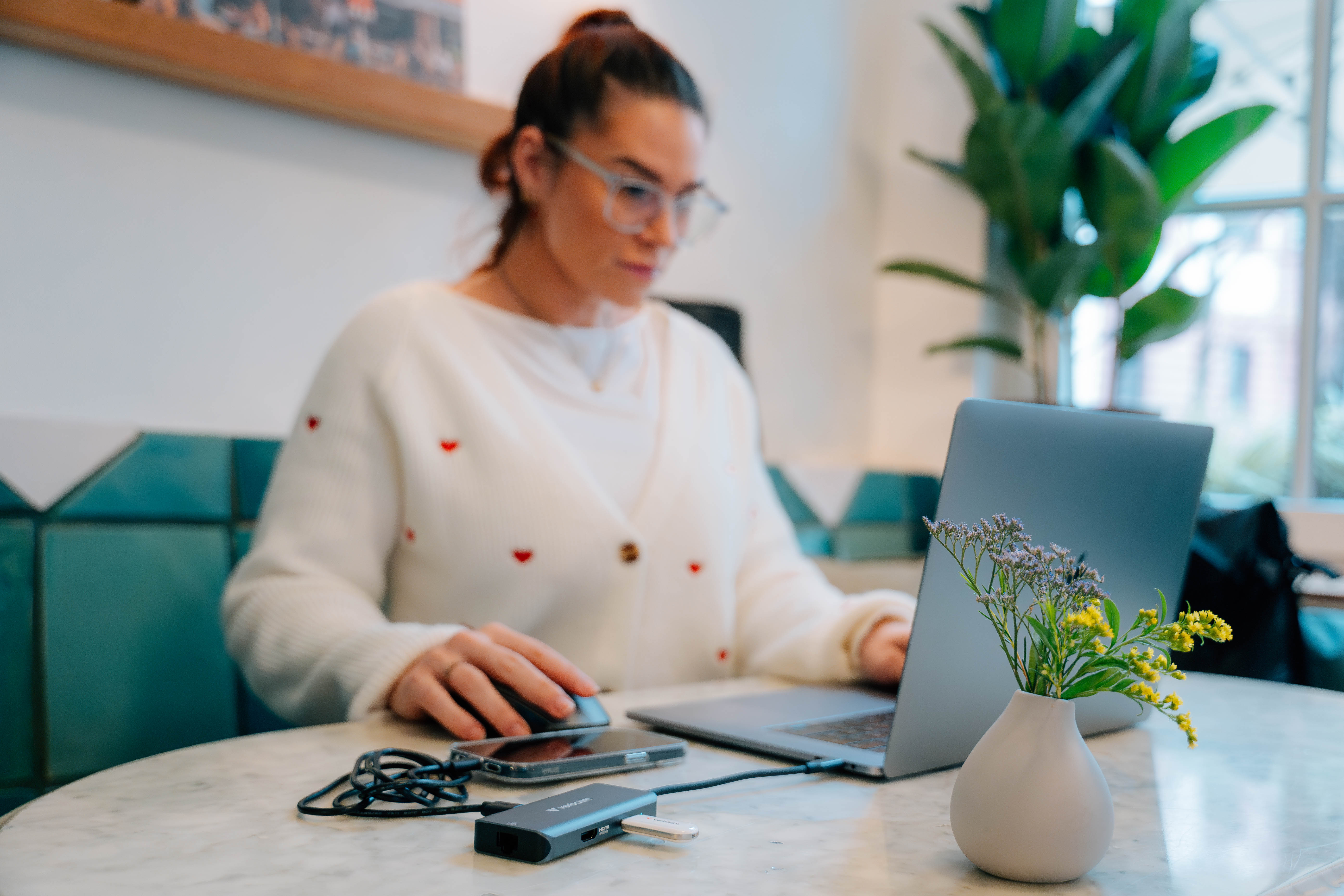 Woman working on laptop with mouse. Flowers in a vase. Phone and USB hub on the table. White sweater.