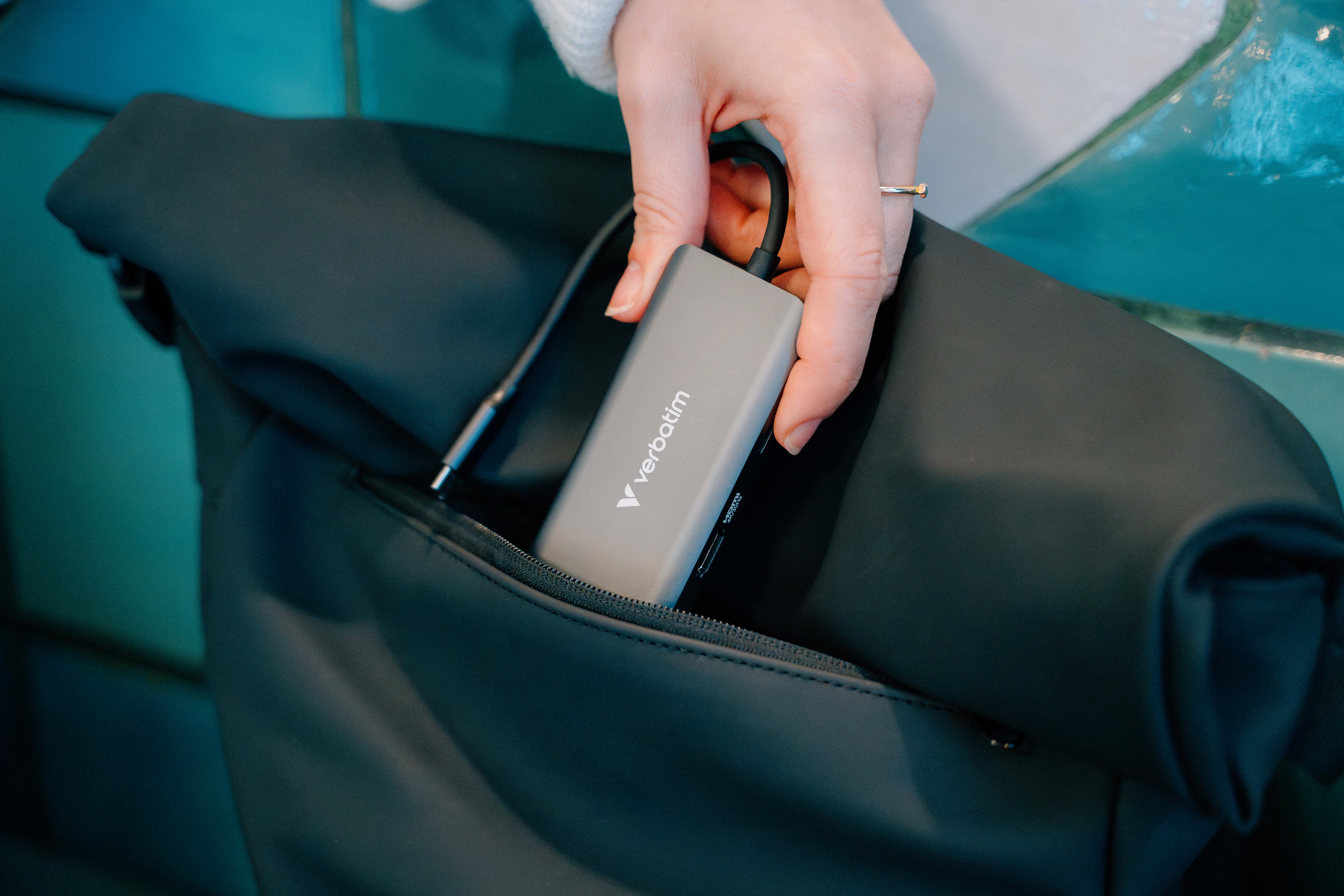 A hand puts a gray Verbatim hub into a black bag pocket. The background is teal.
