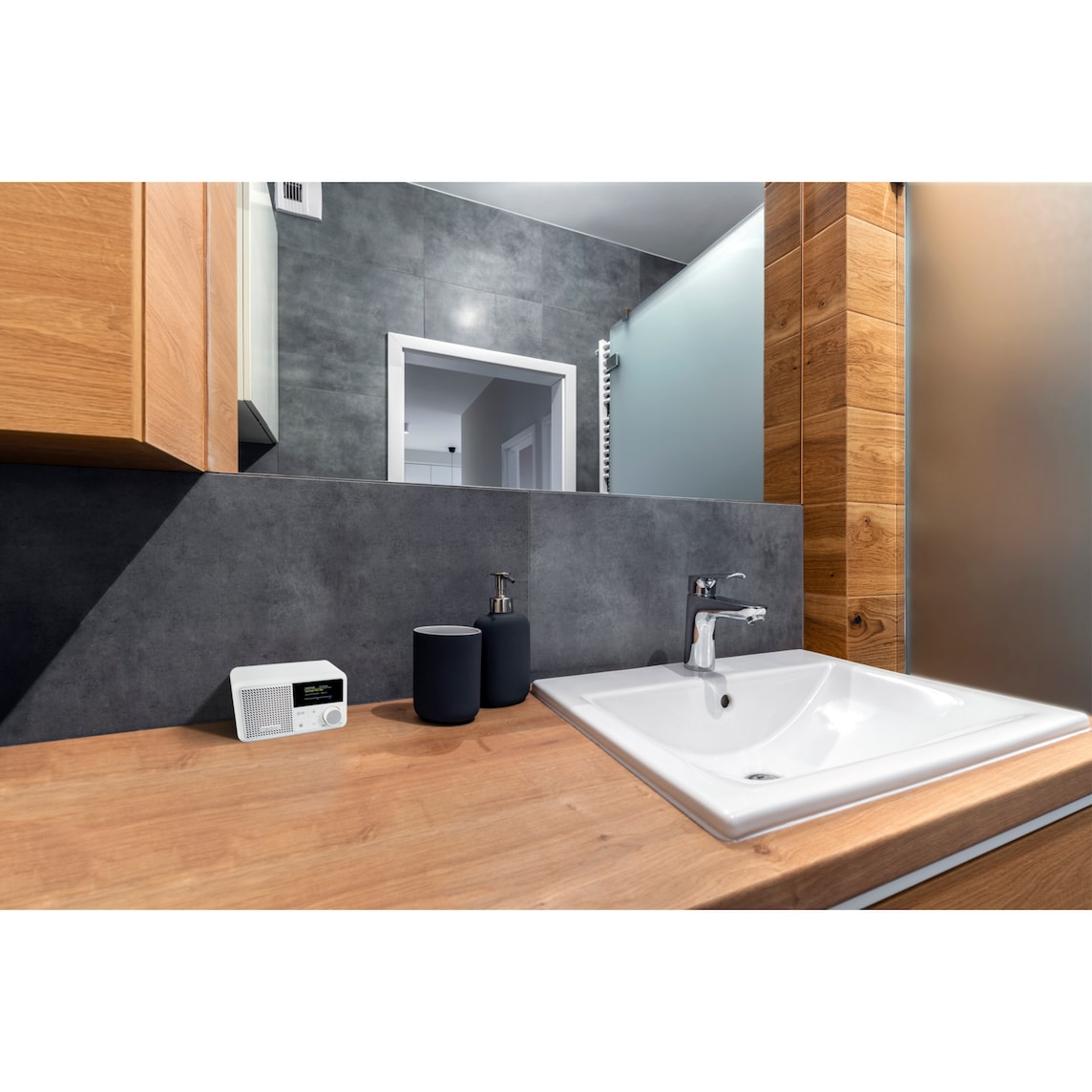A bathroom scene with a wooden countertop, a white sink, a mirror, a radio, and dark blue soap dispenser and cup.