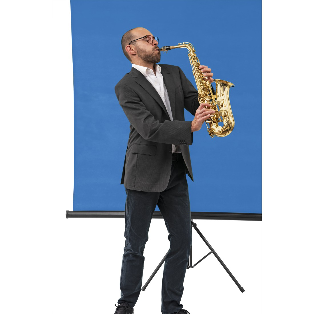 Ein Mann im Anzug spielt ein goldenes Saxophon vor blauem Hintergrund. Er trägt eine Brille und bläst in das Instrument.