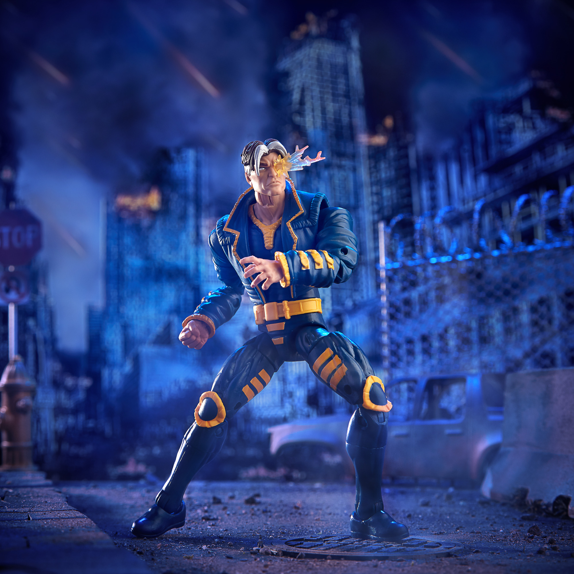 Action figure di un supereroe in tuta blu e gialla, occhio che emette energia, in una città notturna.