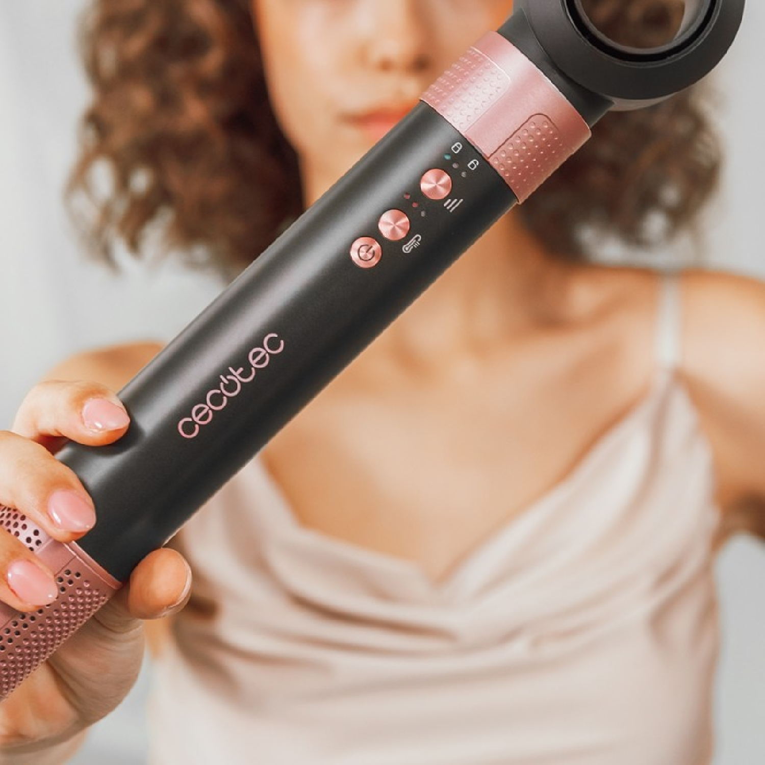 Una donna tiene in mano un arricciacapelli nero e oro rosa. Lo strumento ha dei pulsanti e il marchio 'cecotec' è visibile. La donna indossa un top color crema.