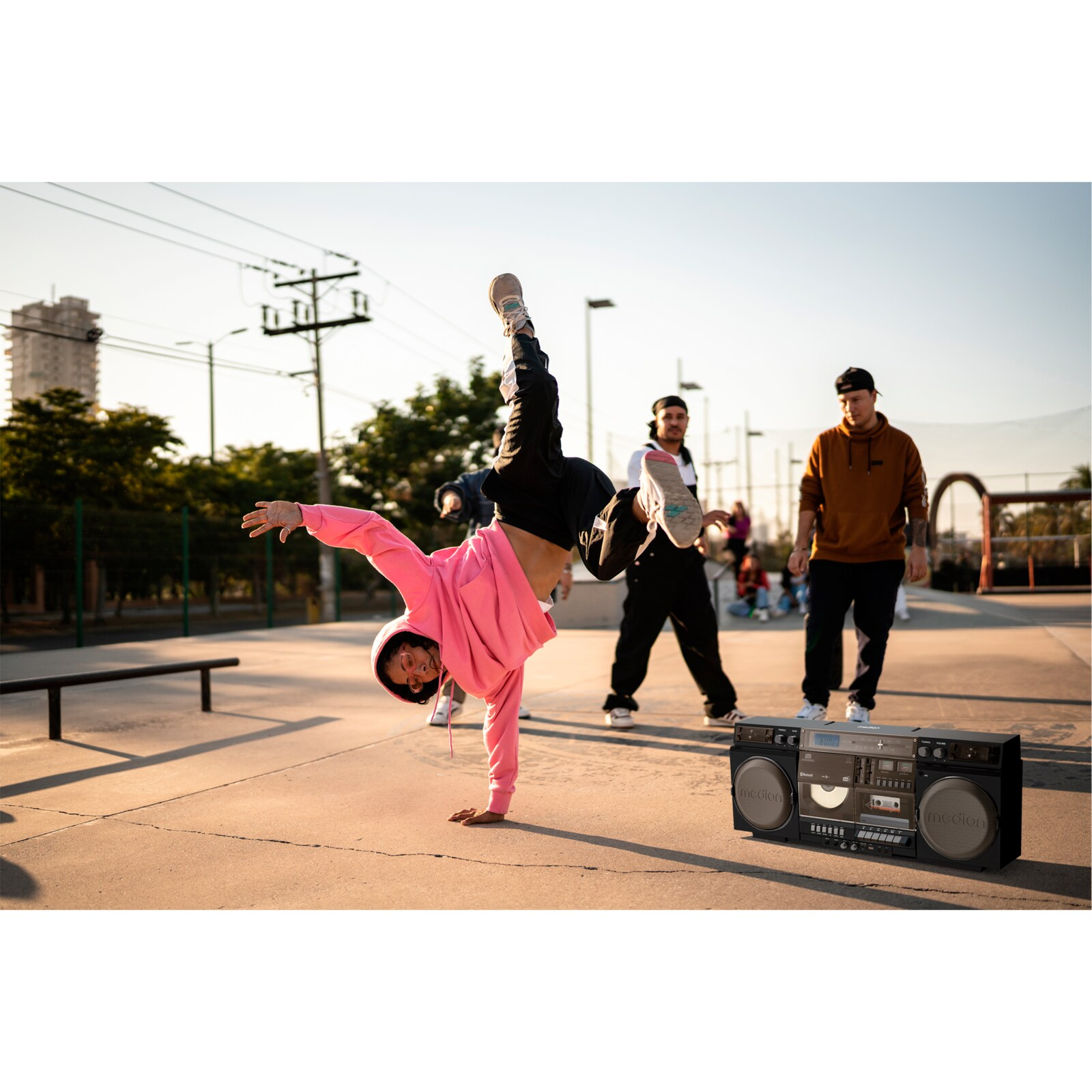 Breakdancer robi stanie na rękach. Dwóch innych stoi za nim z boomboxem. Tłem jest słoneczny skatepark.