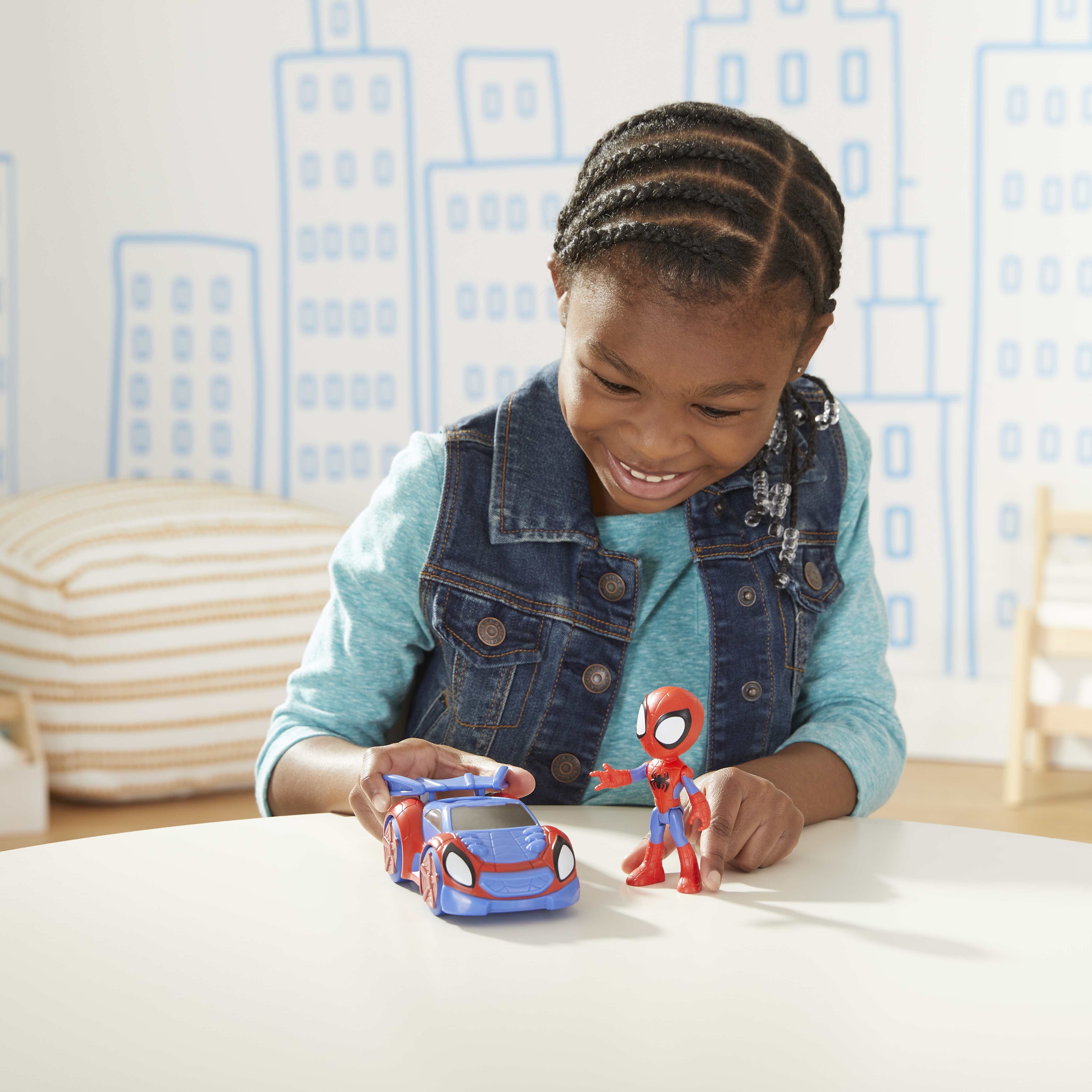 Dziewczyna uśmiecha się do zabawek Spider-Mana. Niebieski samochodzik stoi obok Spidermana. Zarys miasta na ścianie.