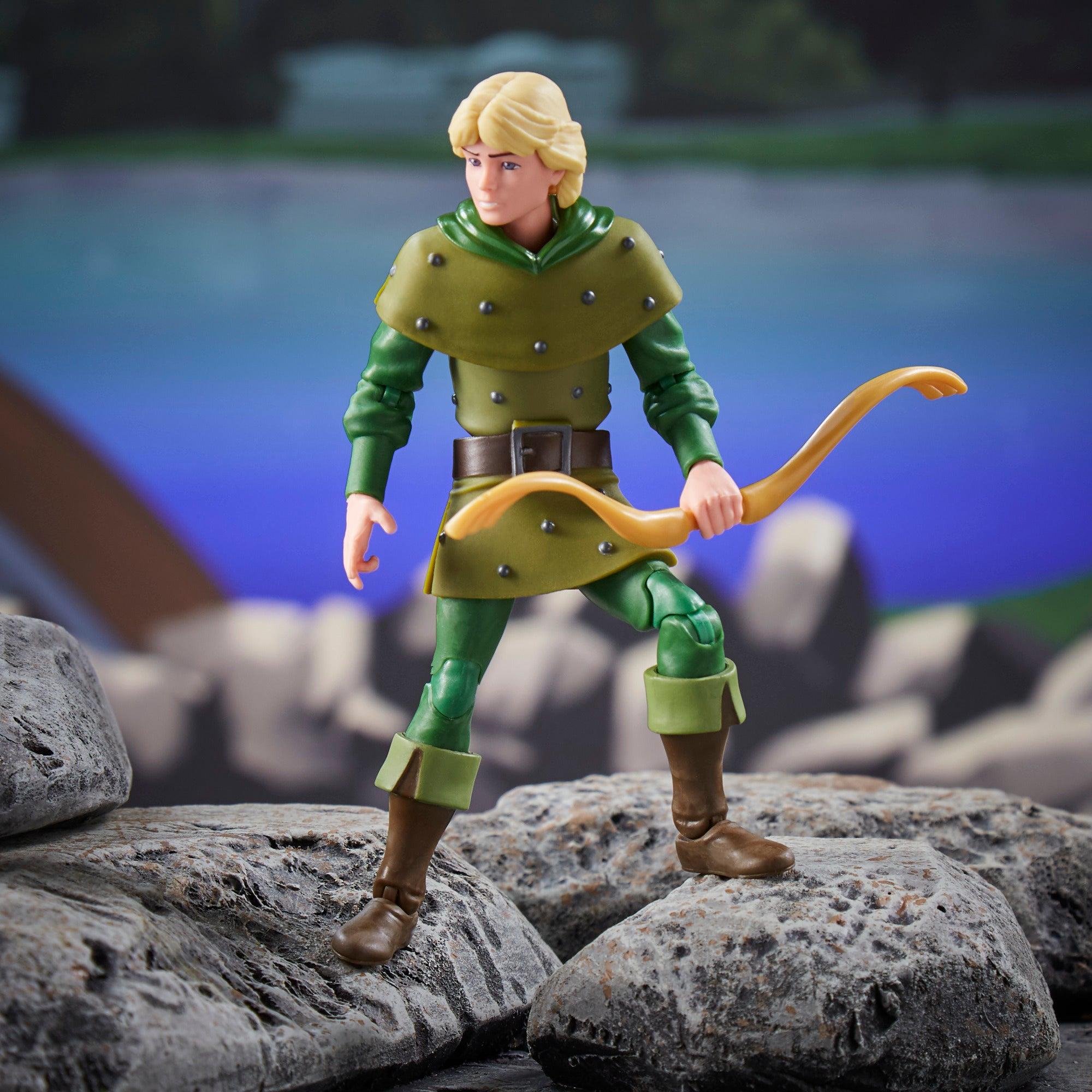 Action figure di un uomo in abito verde con capelli biondi, che tiene un arco, in piedi sulle rocce. Lo sfondo mostra un paesaggio sfocato.