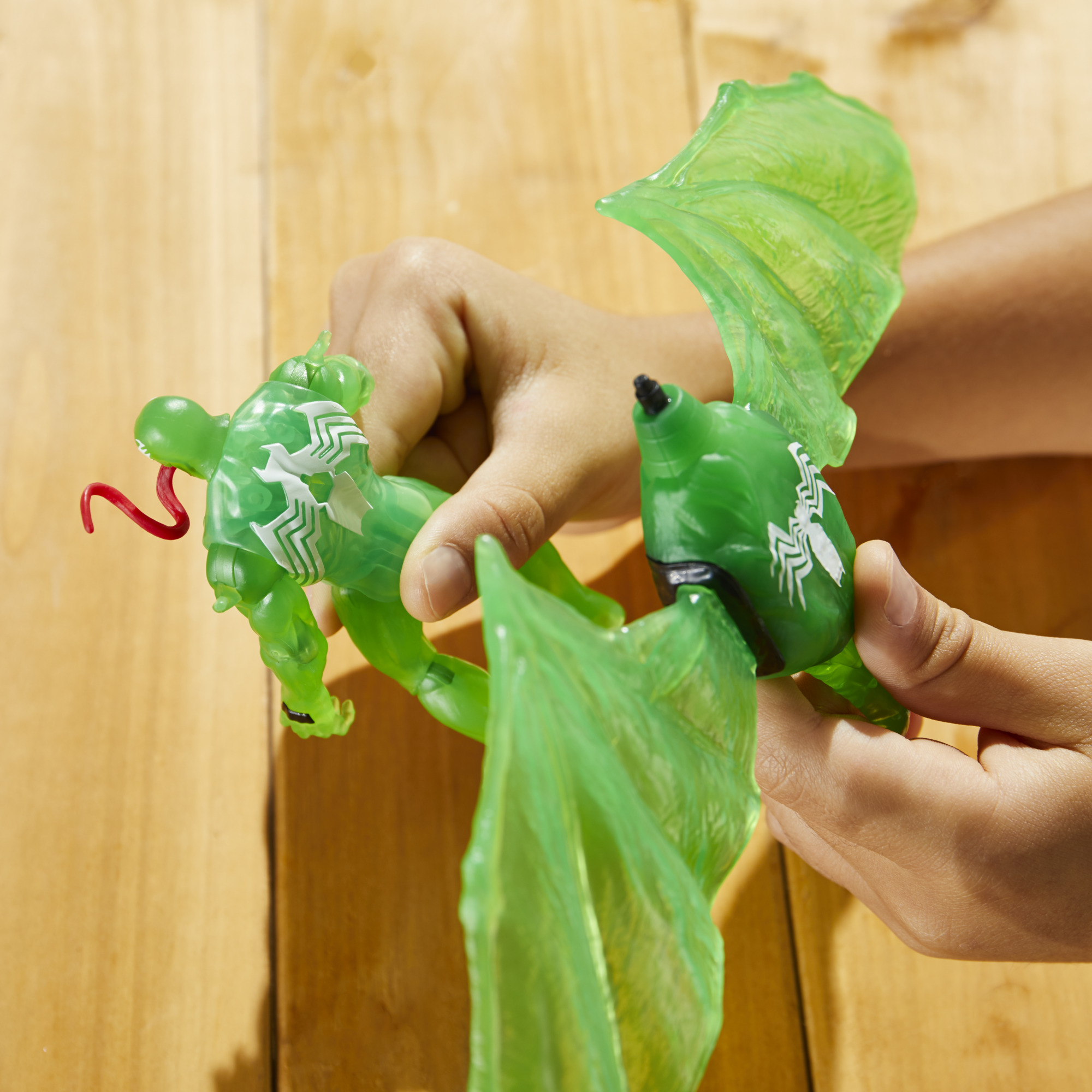 Una persona tiene in mano una action figure verde di Venom con ali, una lingua rossa e un simbolo di ragno bianco su sfondo di legno.