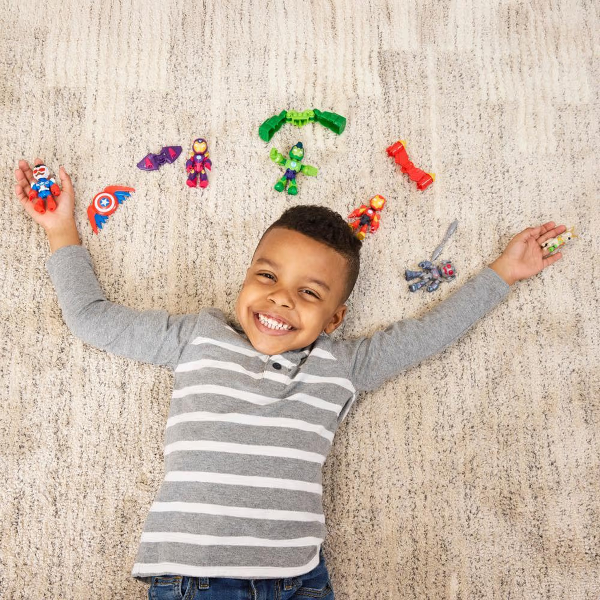 Un niño sonriente yace en una alfombra con superhéroes de juguete a su alrededor. Viste una camisa gris a rayas y vaqueros.