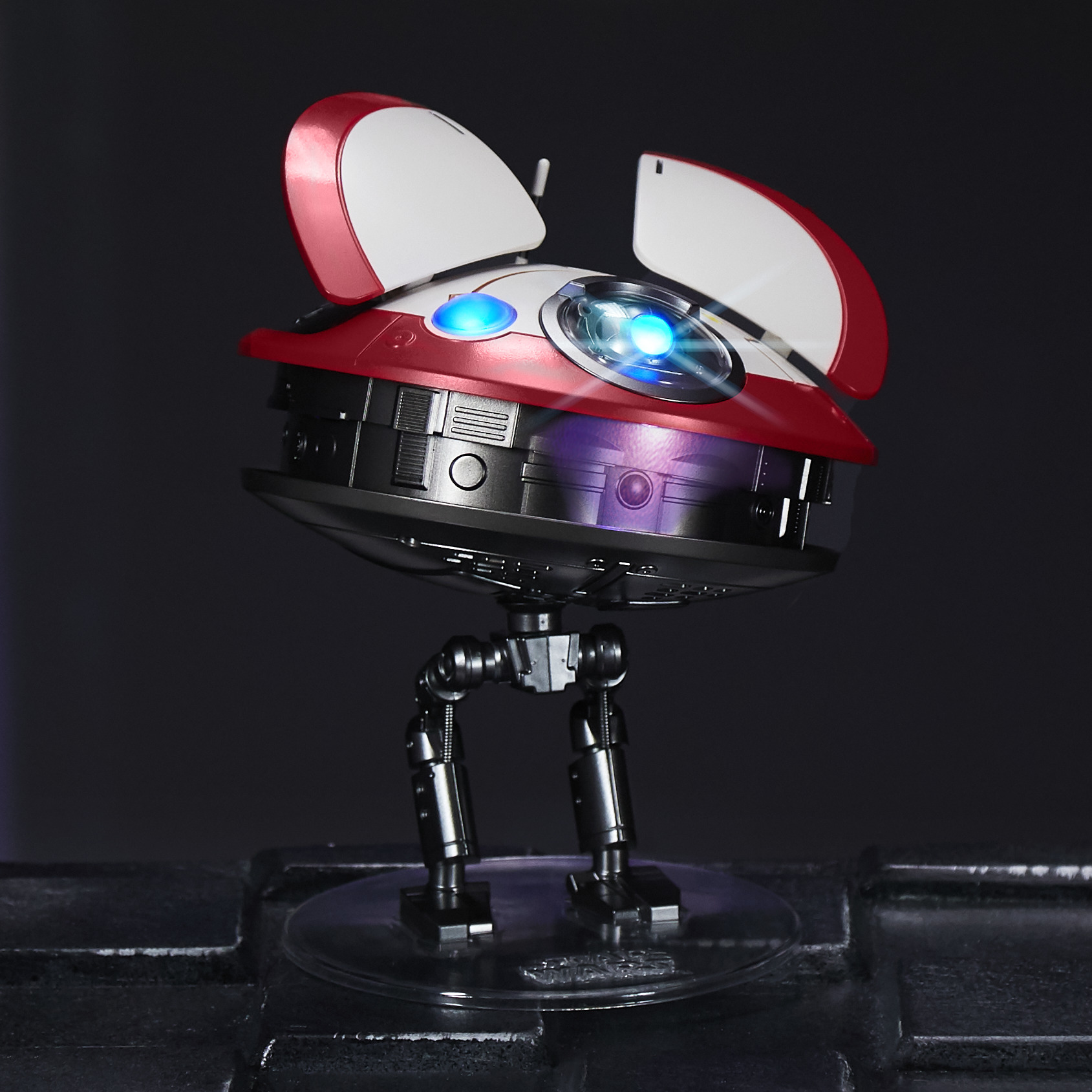 Zabawka robota z czerwoną i białą głową, niebieskimi światłami i srebrnymi nogami, na czarnej powierzchni.