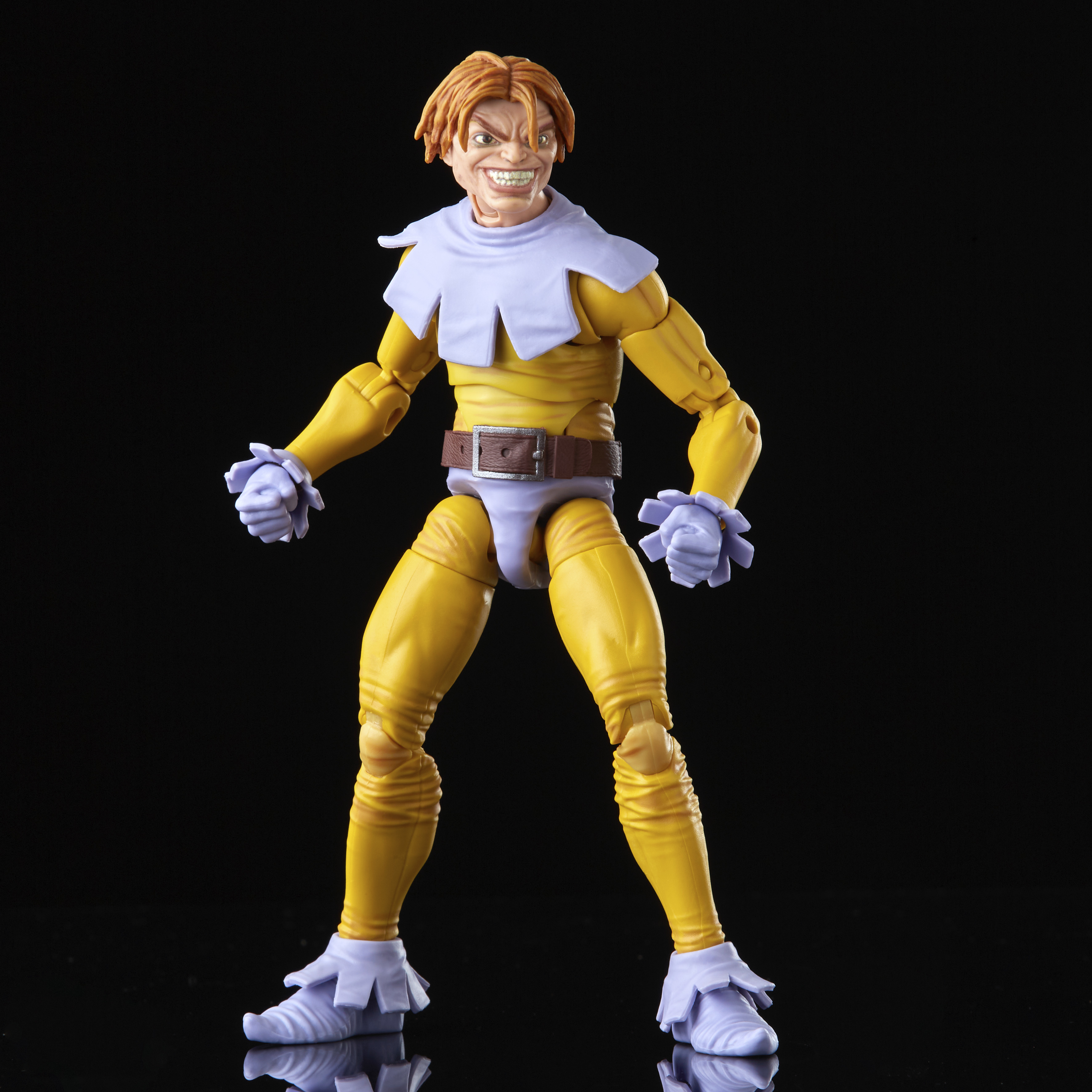 Una action figure gialla con un sorriso minaccioso si erge su uno sfondo nero. Ha colletto e polsini bianchi.