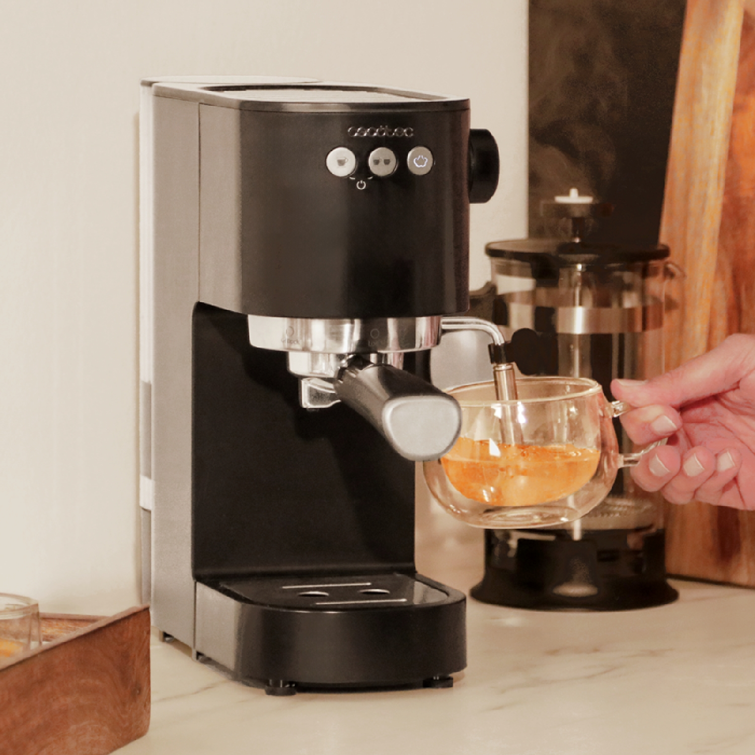 Eine schwarze Espressomaschine gibt Kaffee in eine Glastasse, die von einer Hand gehalten wird. Im Hintergrund sind eine French Press und ein Holzbrett zu sehen.