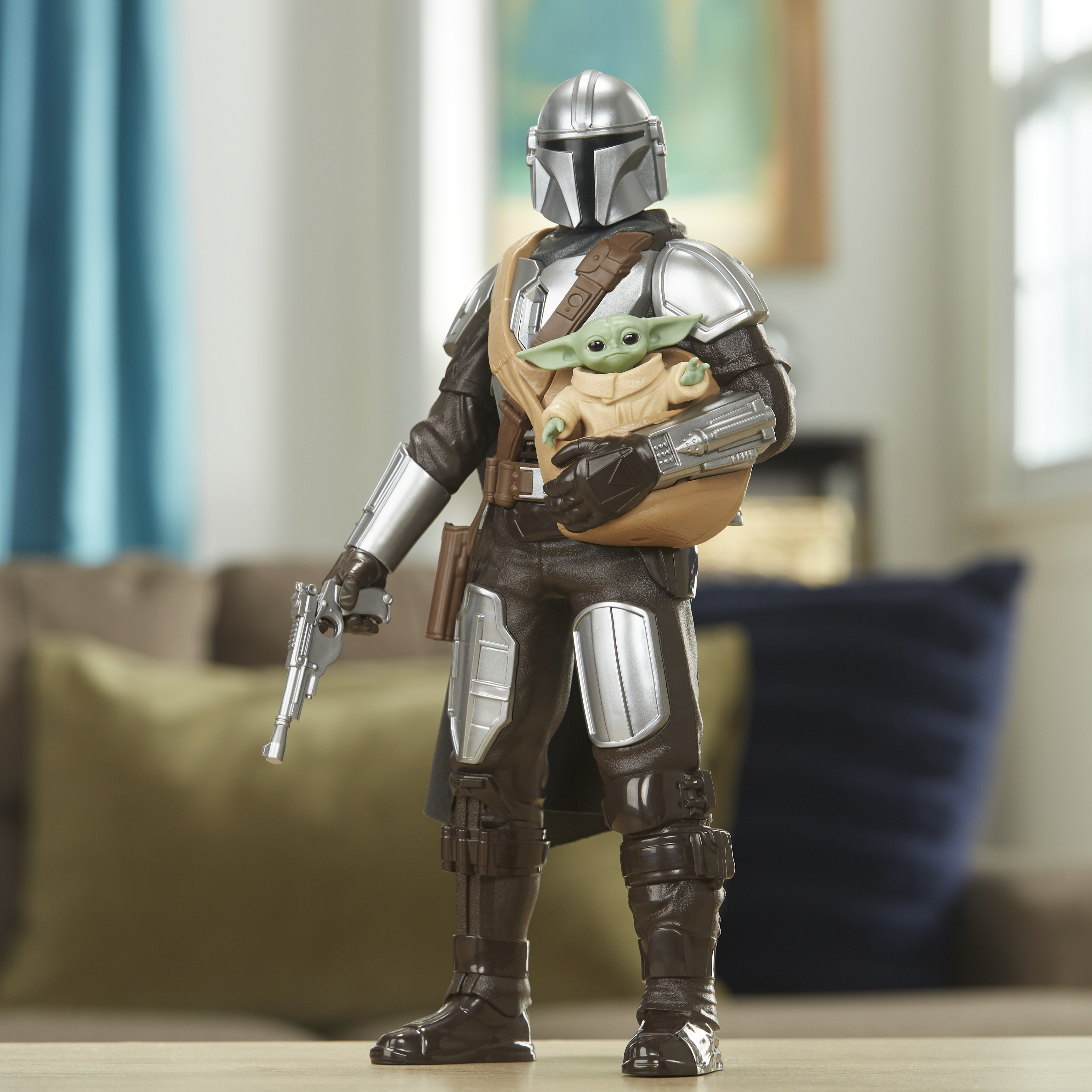 La figura del Mandaloriano tiene in mano Baby Yoda. È su un tavolo. Tenda blu e divano marrone sullo sfondo.