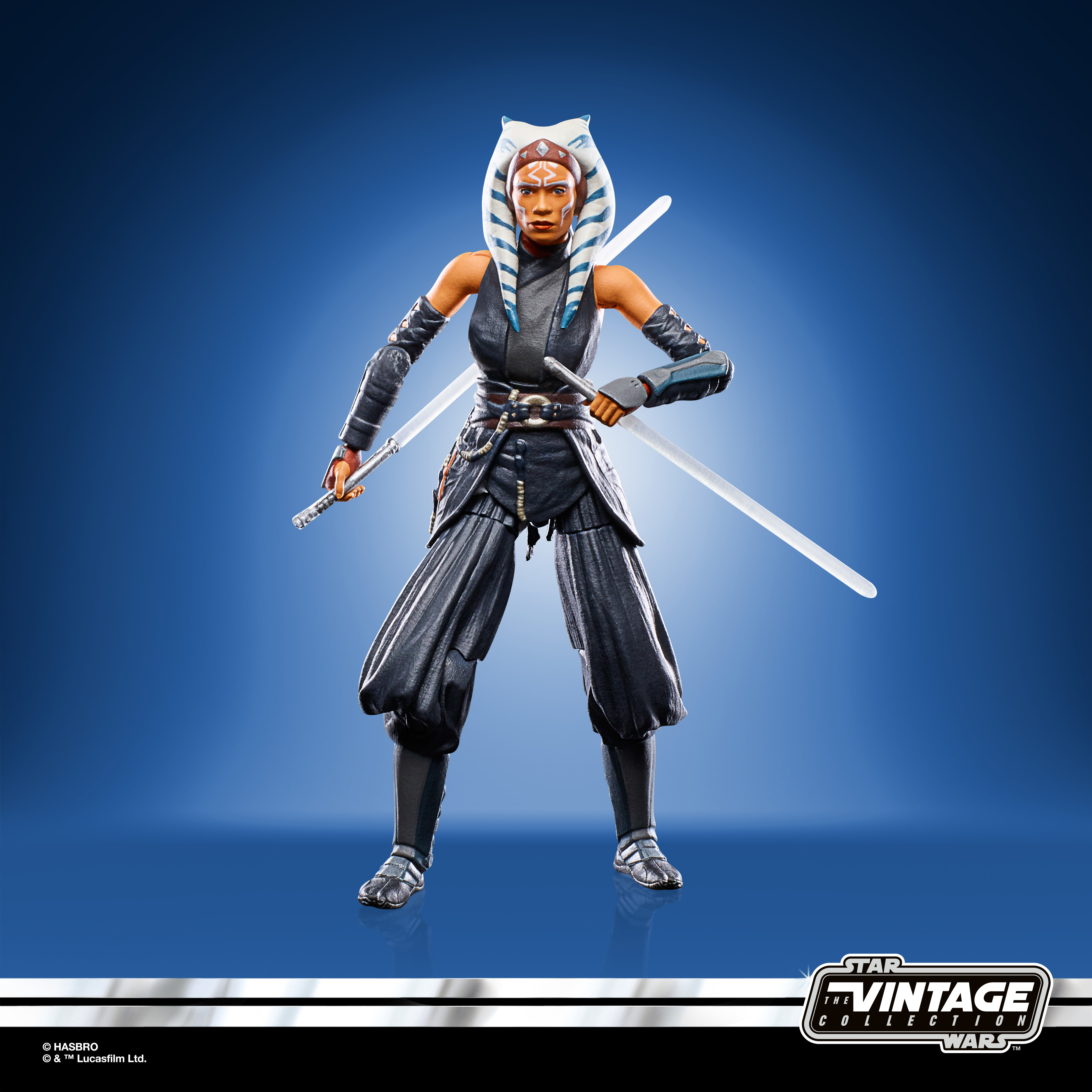 Figurka akcji Star Wars Ahsoka Tano z dwoma mieczami świetlnymi, w szarych i czarnych ubraniach, na niebieskim tle.