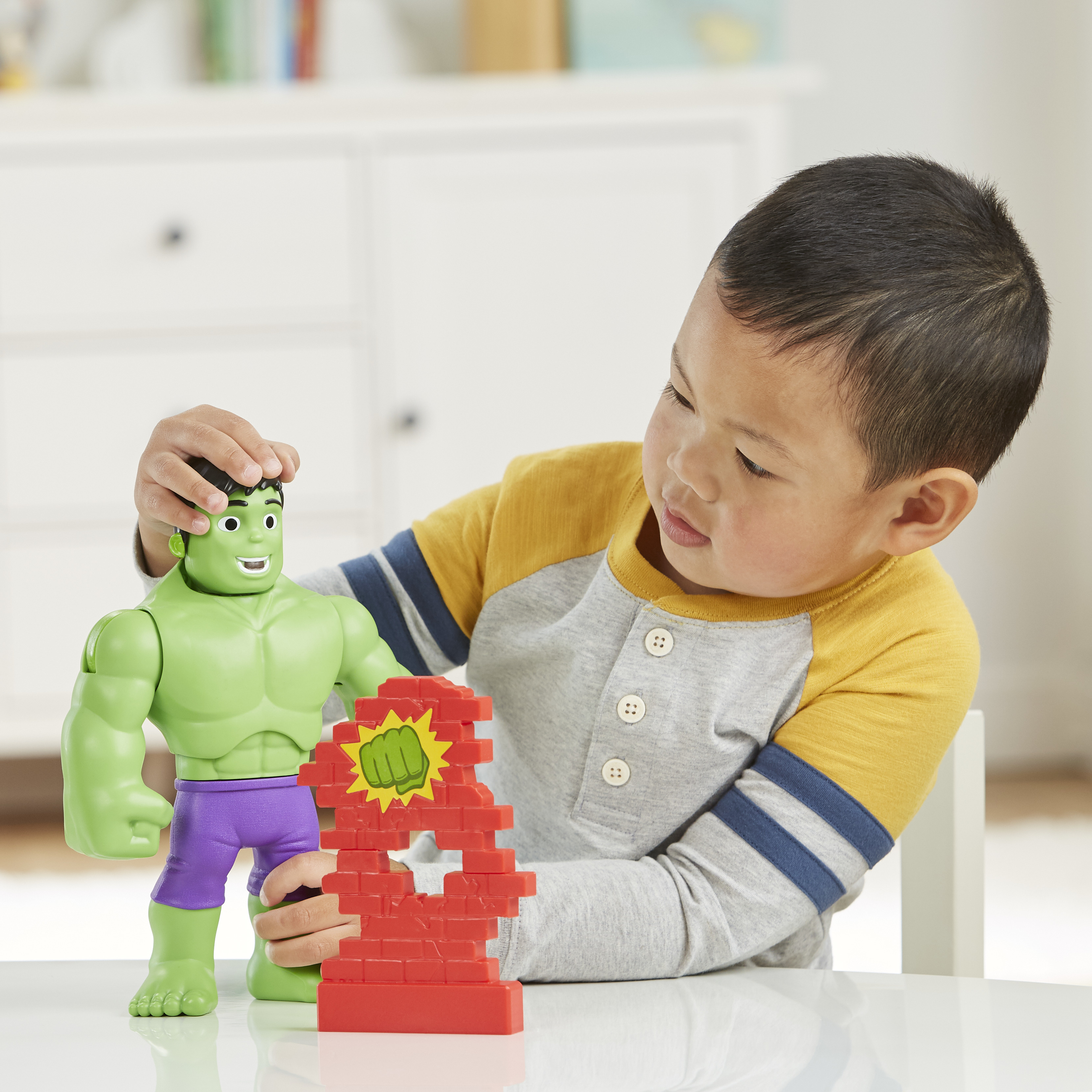 Un bambino gioca con un giocattolo di Hulk e un giocattolo a muro di mattoni. Il bambino indossa una camicia gialla e grigia.