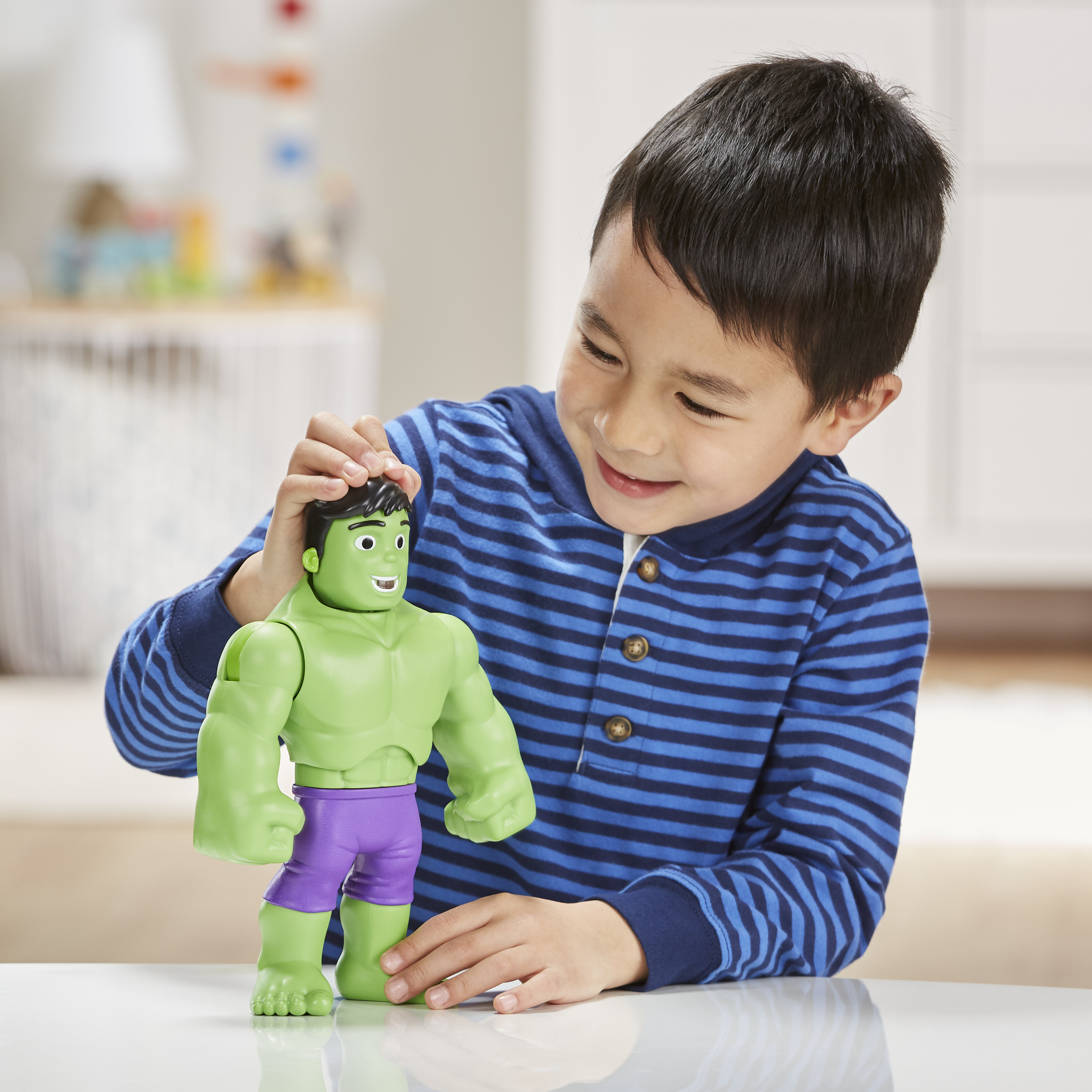 Un ragazzo sorride, tenendo in mano un giocattolo di Hulk verde con pantaloncini viola. Il ragazzo indossa una camicia a righe blu.