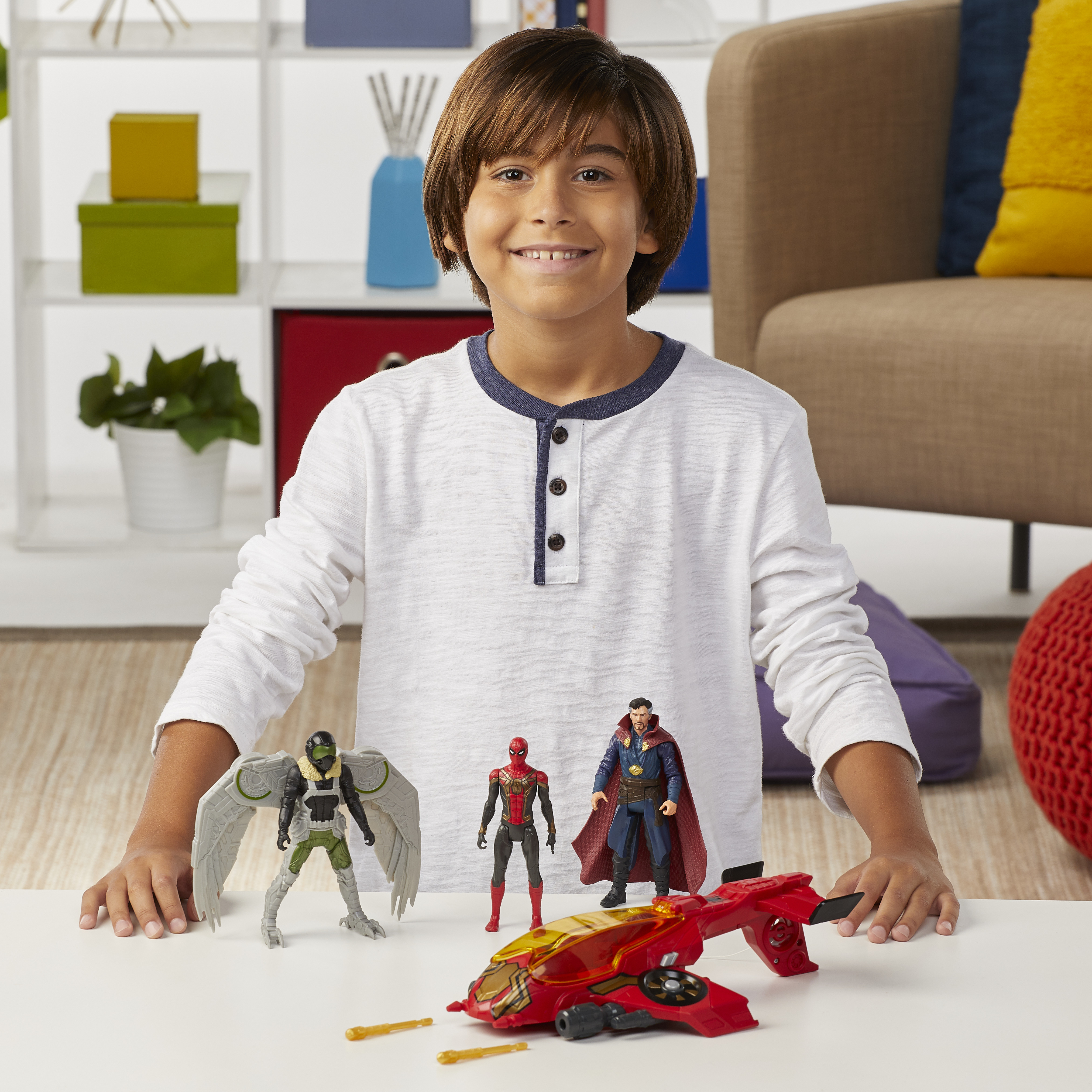 Un ragazzo sorride, presentando figure di Spider-Man, Vulture, Doctor Strange e un jet rosso su un tavolo bianco.