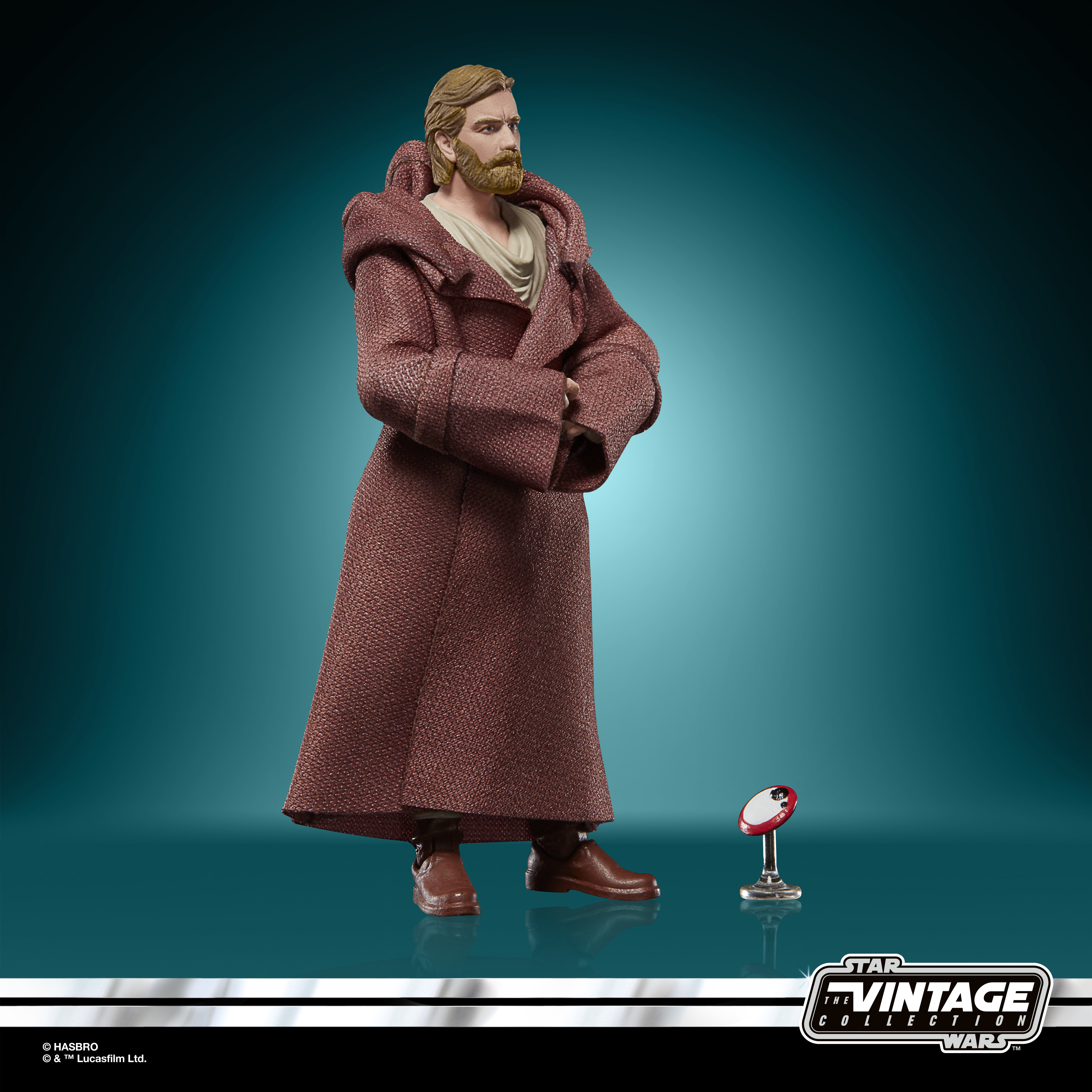 Figura de Obi-Wan Kenobi con abrigo marrón. Está frente a un fondo turquesa con un pequeño objeto rojo y blanco.