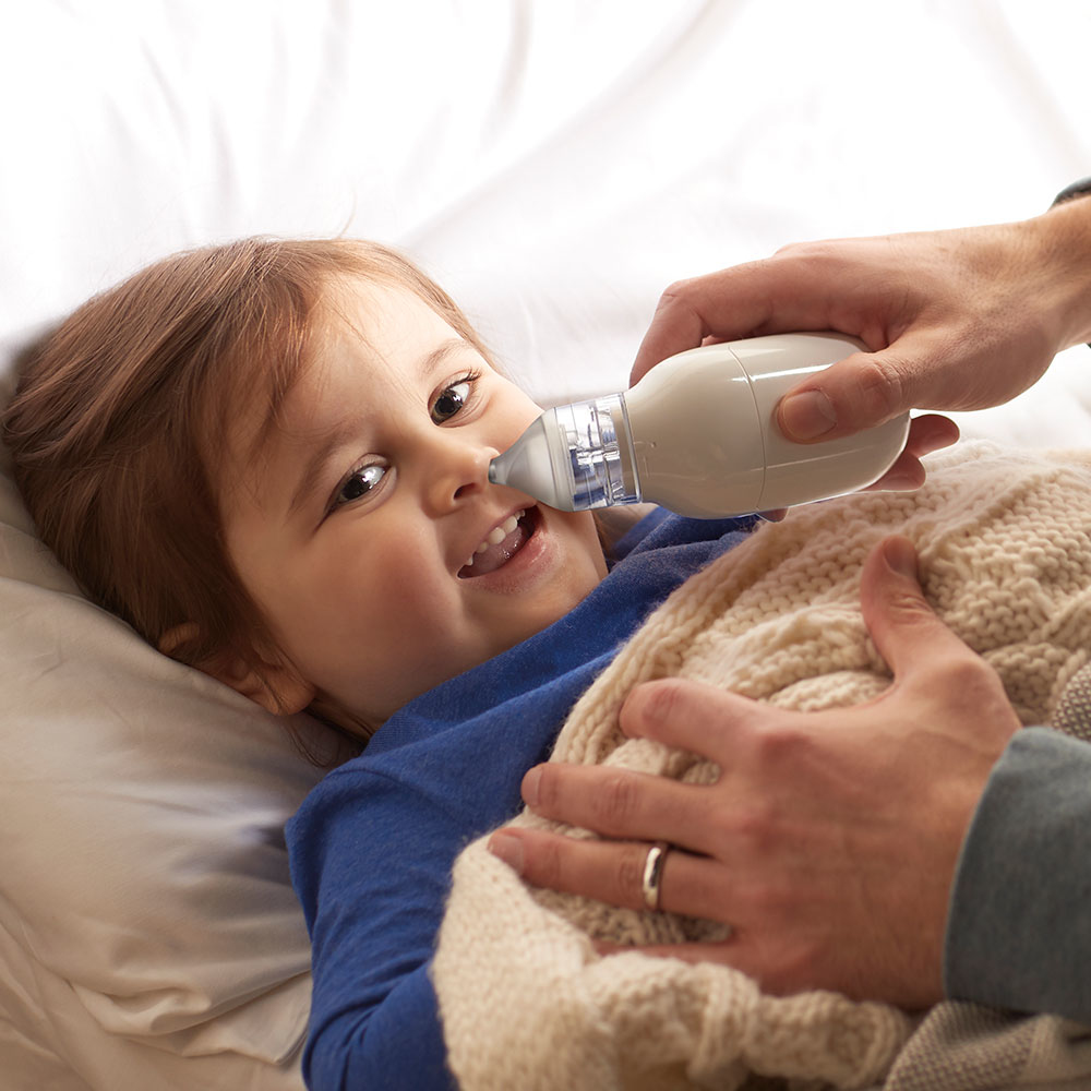 Un niño acostado, le limpian la nariz con un aspirador nasal blanco. Una mano sostiene el dispositivo y otra al niño.