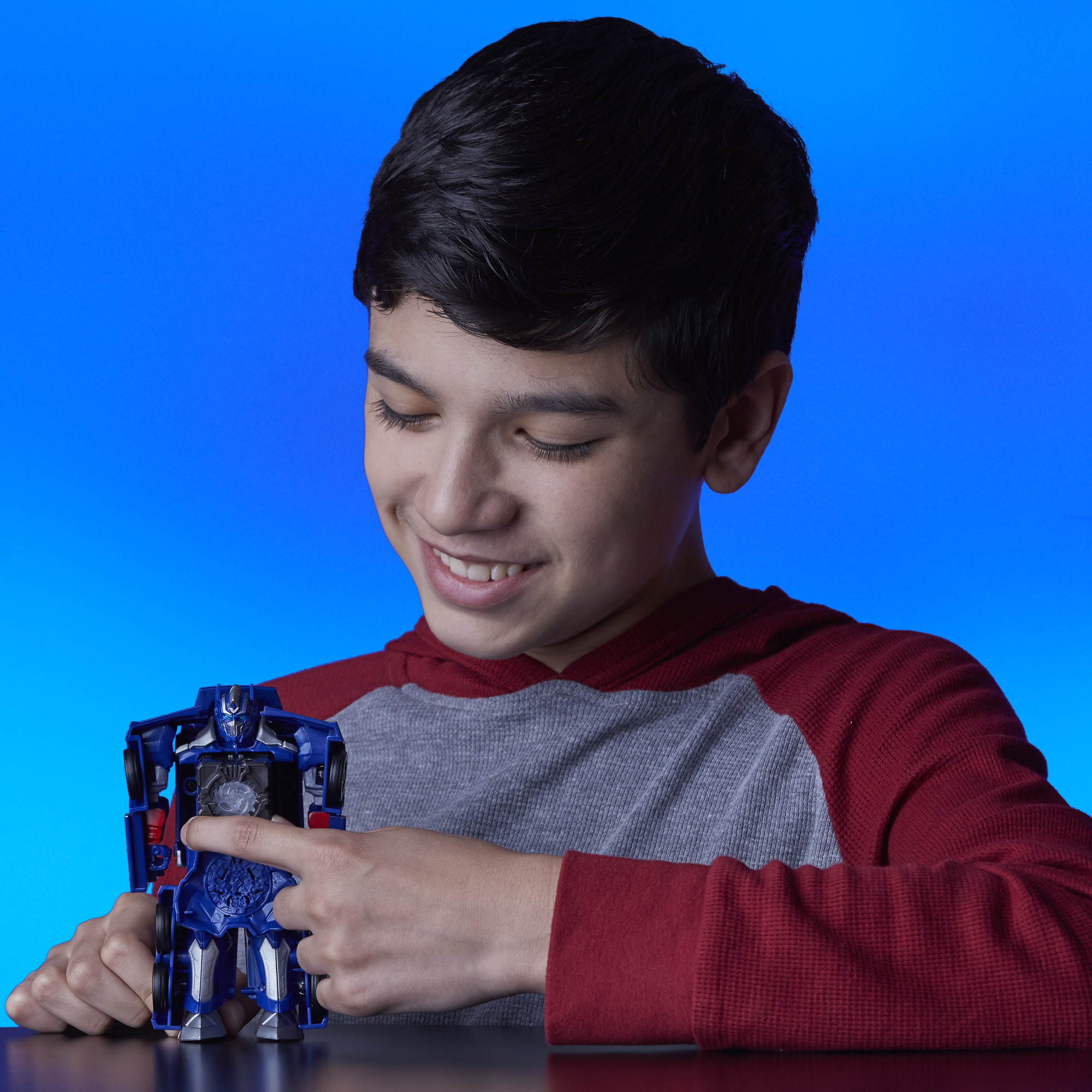 Un ragazzo sorride, guardando un giocattolo Transformer blu. Indossa una camicia rossa e grigia. Lo sfondo è blu.