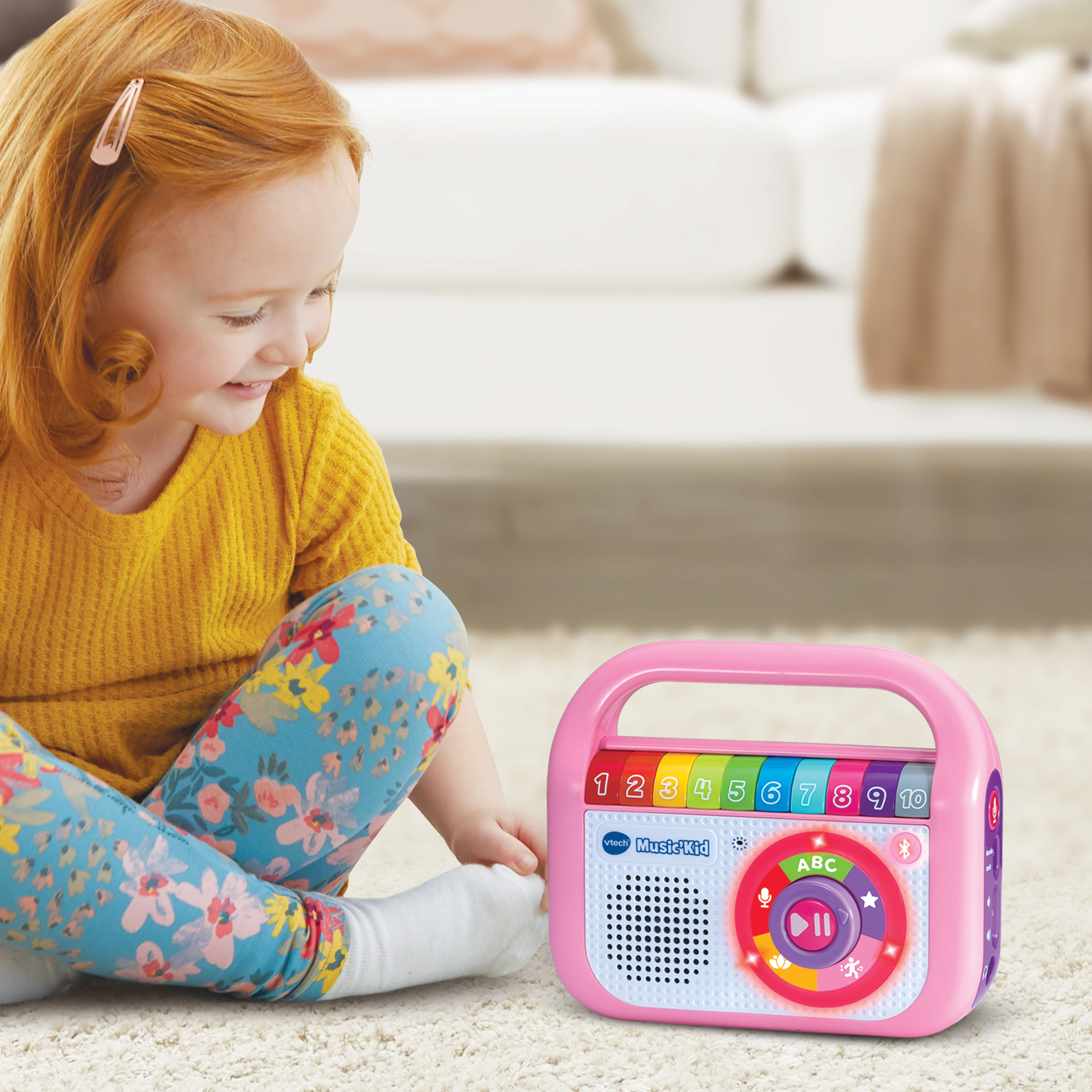 Ein Mädchen mit roten Haaren lächelt einen pinkfarbenen Musikplayer an. Der Player hat bunte Knöpfe und Zahlen. Sie trägt ein gelbes Hemd und Blumenleggings.