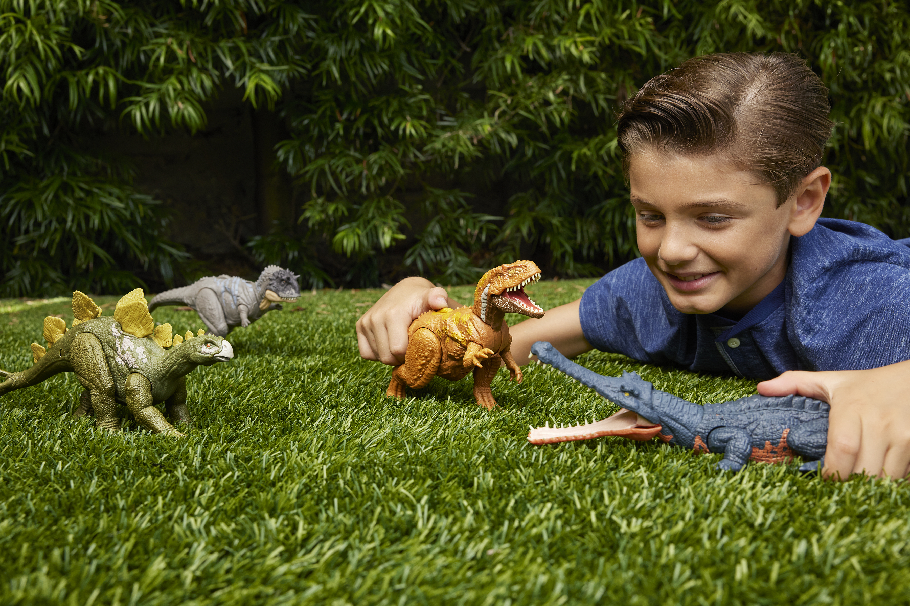 Un niño sonríe, jugando con juguetes de dinosaurios sobre hierba. Los dinosaurios son marrones, grises y verdes. Follaje verde al fondo.