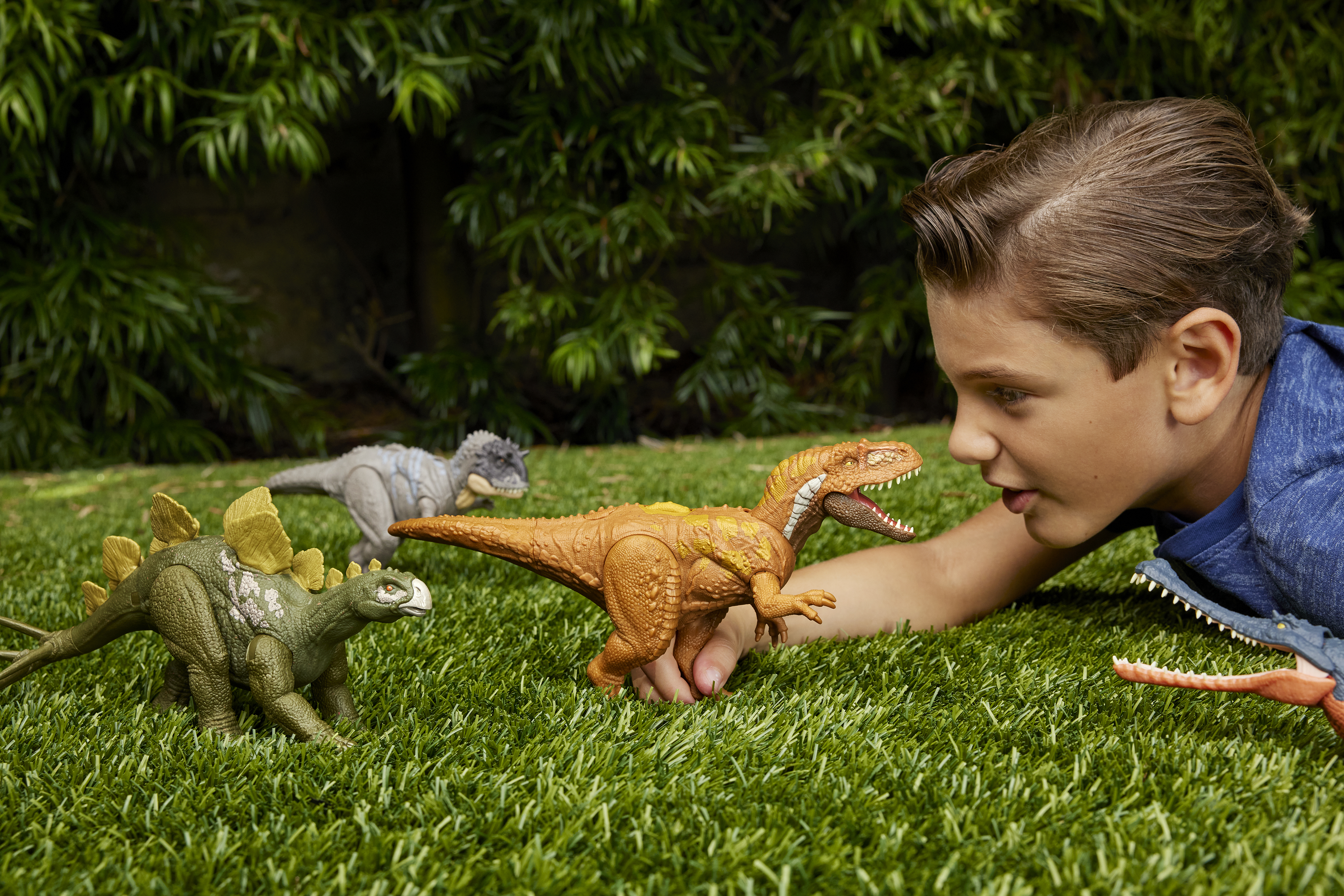 Un niño mira juguetes de dinosaurios sobre hierba. Los dinosaurios son marrones, verdes y grises. Follaje verde al fondo.
