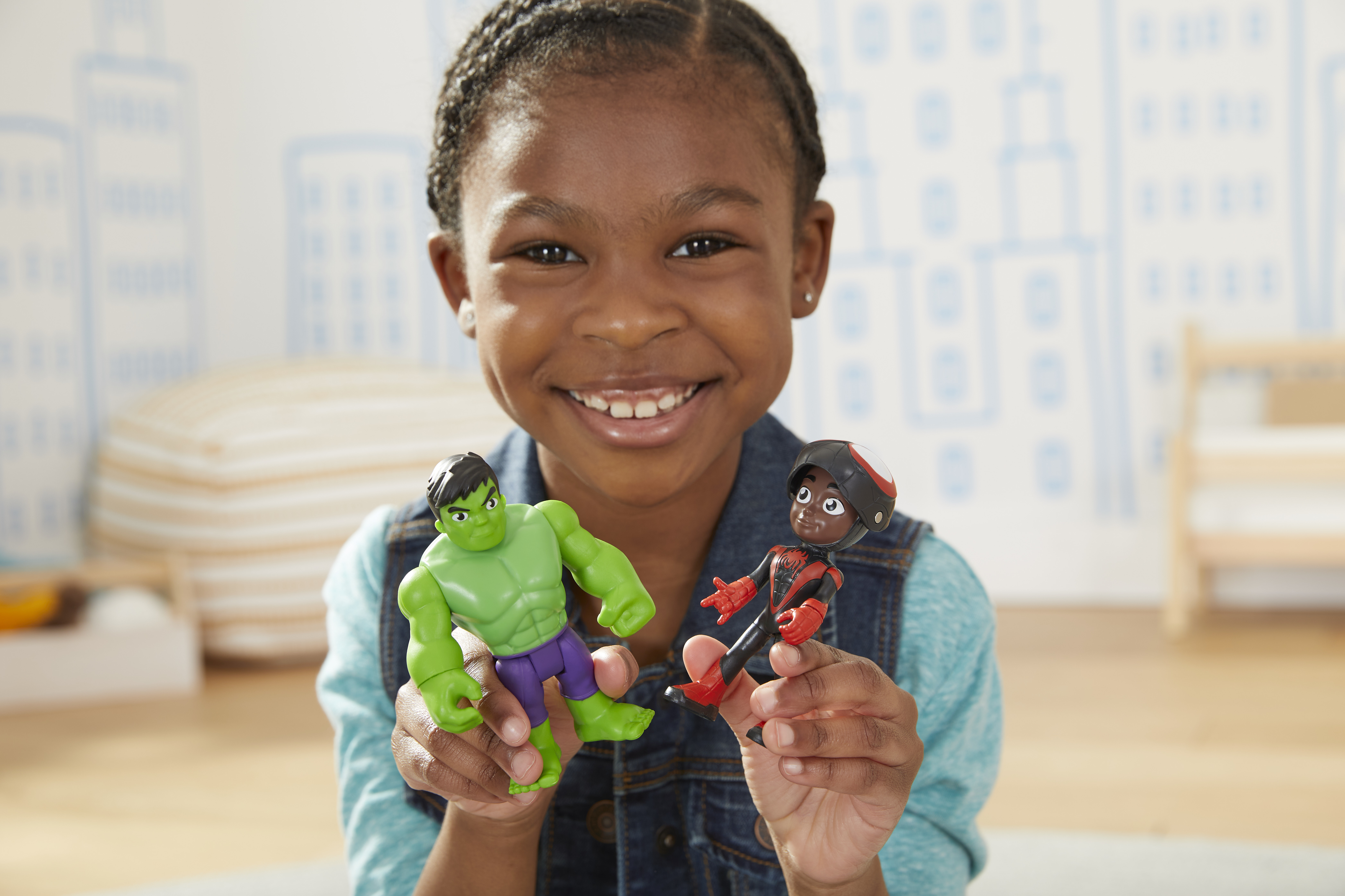 Una ragazza sorridente tiene in mano i personaggi di Hulk e Spider-Man. Indossa un gilet di jeans e una camicia blu. Lo sfondo ha un disegno di città.