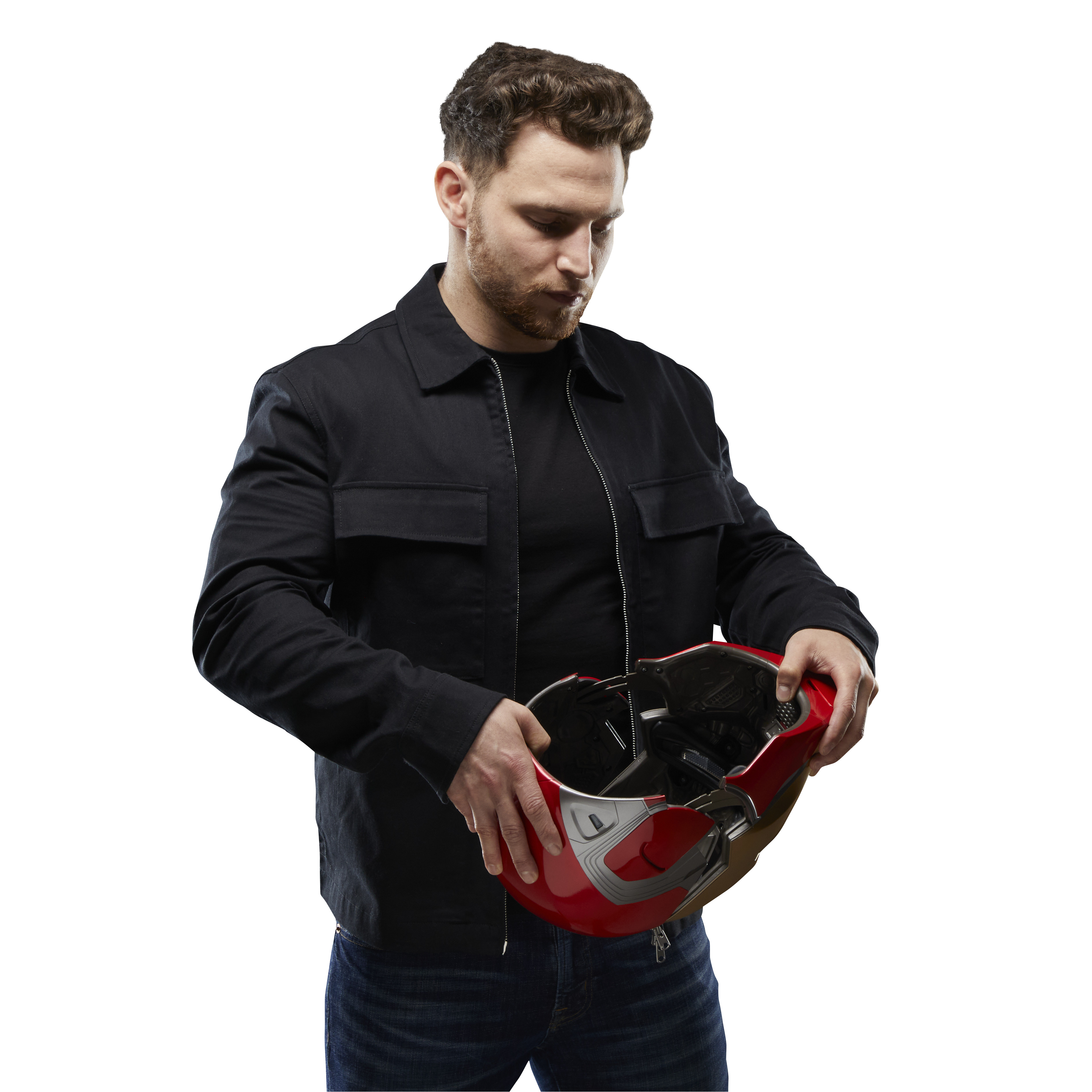 Mężczyzna w czarnej kurtce trzyma czerwony kask ze złotymi detalami. Patrzy w dół. Białe tło.