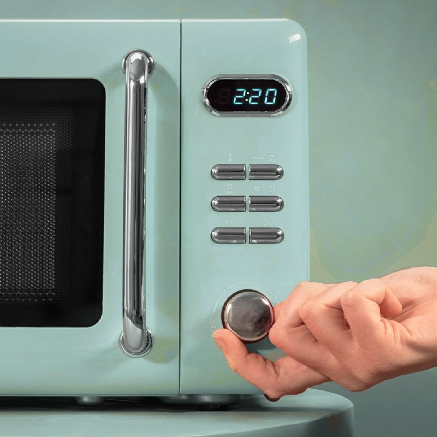 Una mano gira la manopola di un forno a microonde verde menta. Il display mostra 8:20. I pulsanti sono visibili. La maniglia è d'argento.