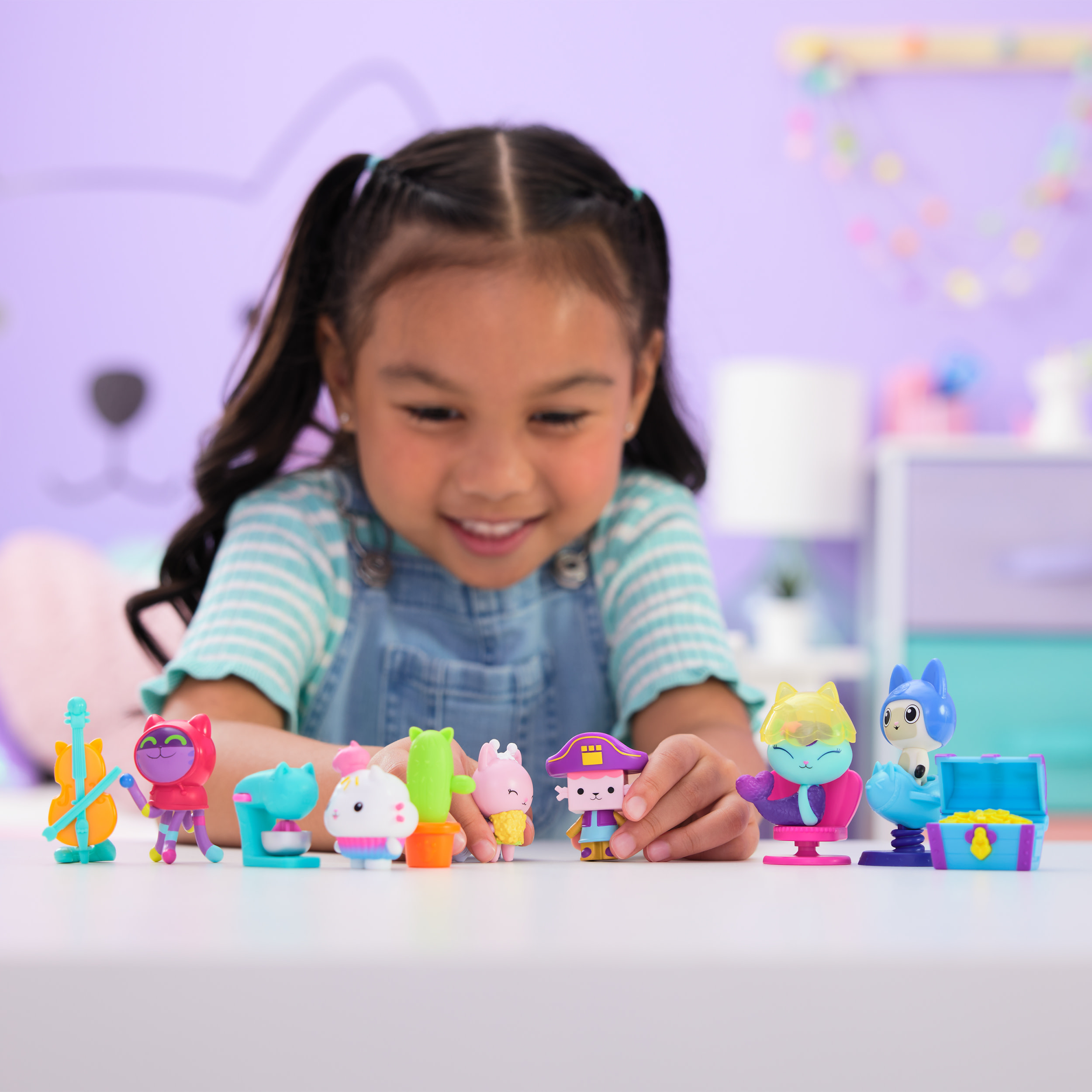 Una niña sonríe a figuras de juguete coloridas sobre una mesa blanca. El fondo es una pared morada con un diseño de gato.