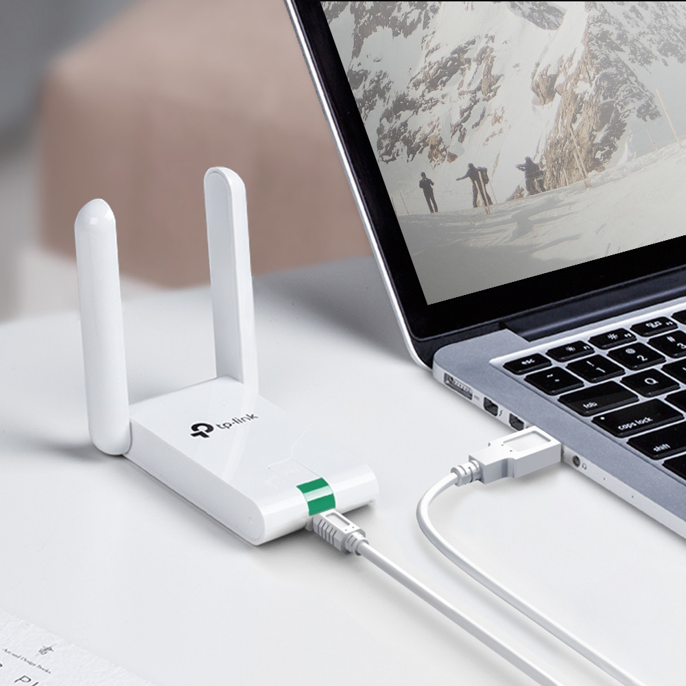 Biały adapter Wi-Fi TP-Link jest podłączony do laptopa. Ekran pokazuje zaśnieżoną górską scenerię. Konfiguracja znajduje się na białym biurku.