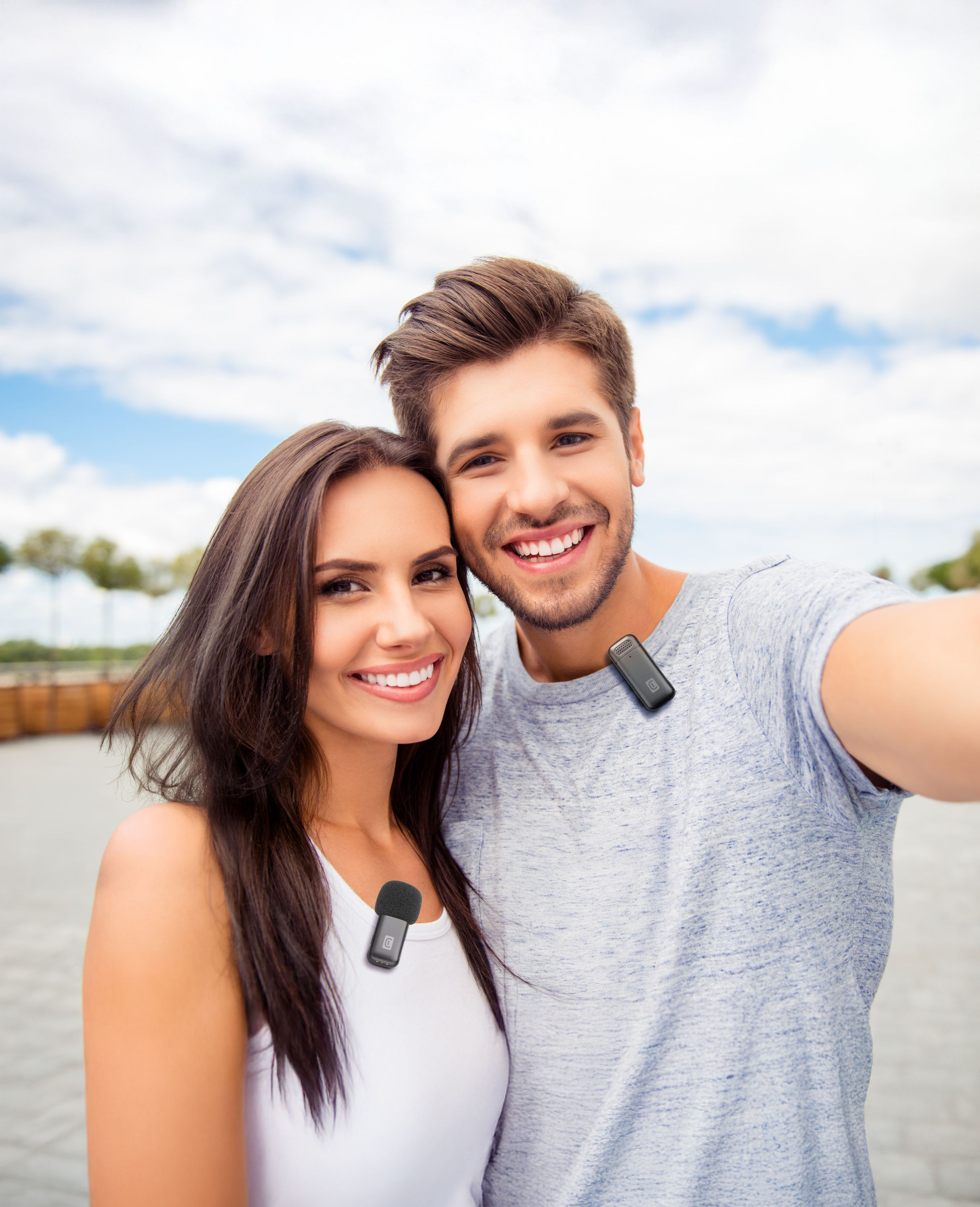 Una coppia sorridente fa un selfie. Indossano microfoni e sono contro un cielo nuvoloso.