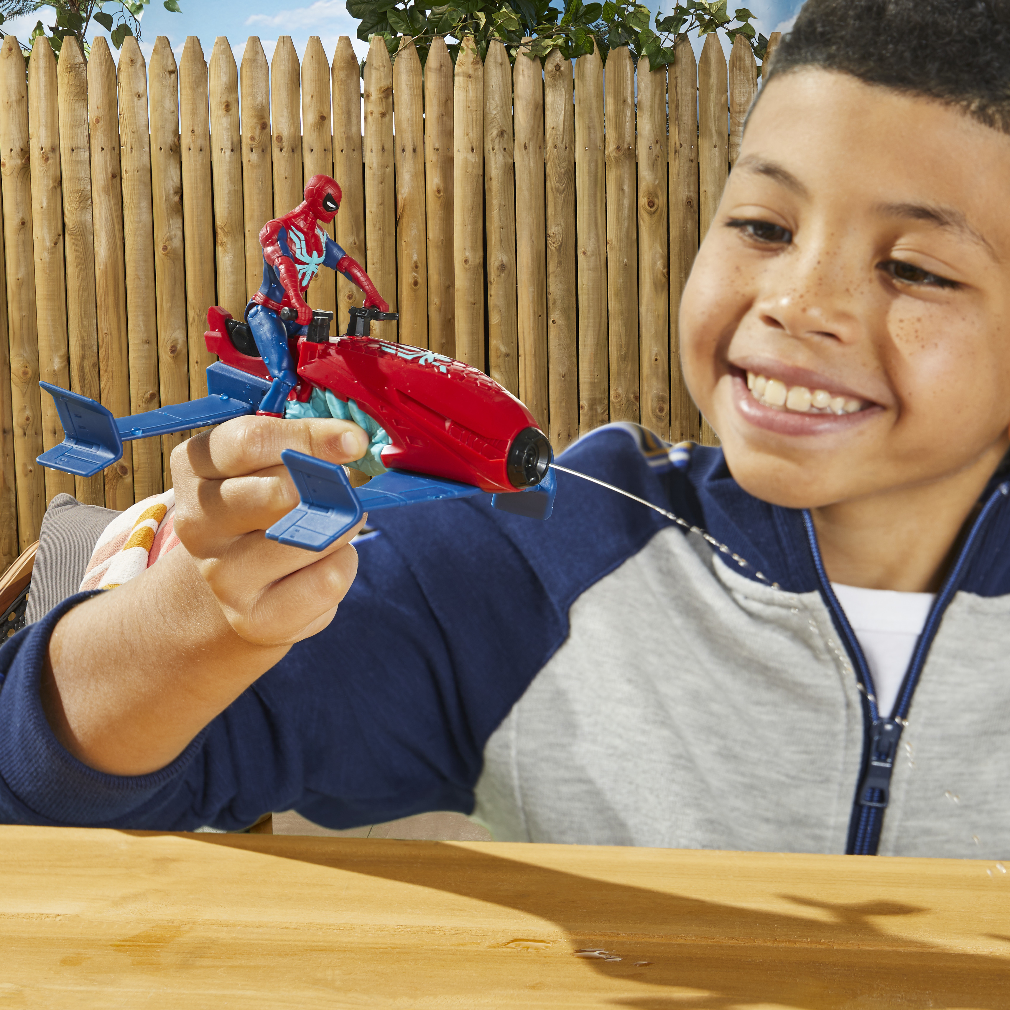 Un ragazzo sorride, tenendo in mano un jet di Spiderman giocattolo. L'acqua spruzza dal jet. Sullo sfondo c'è una recinzione di legno.