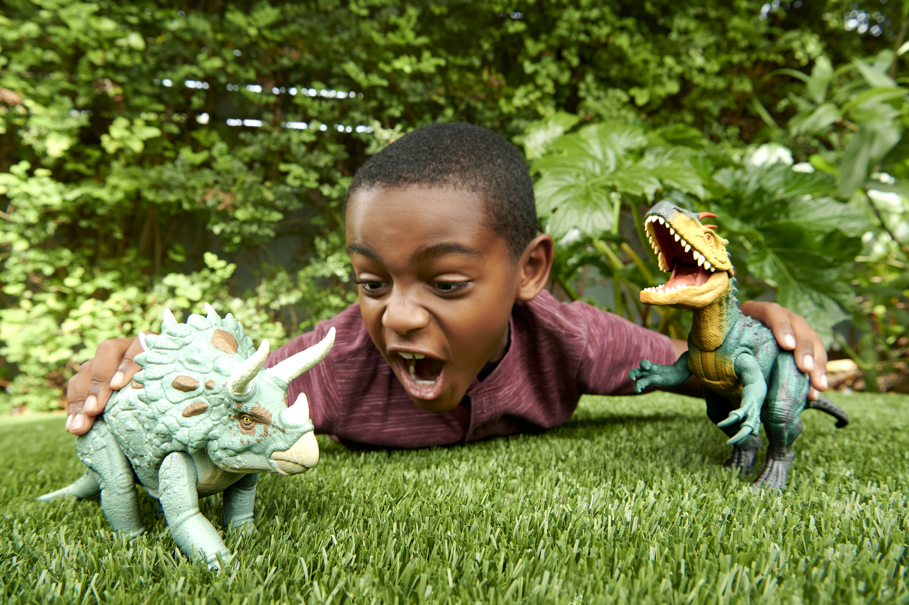Un niño juega con dinosaurios de juguete sobre la hierba. Follaje verde en el fondo.