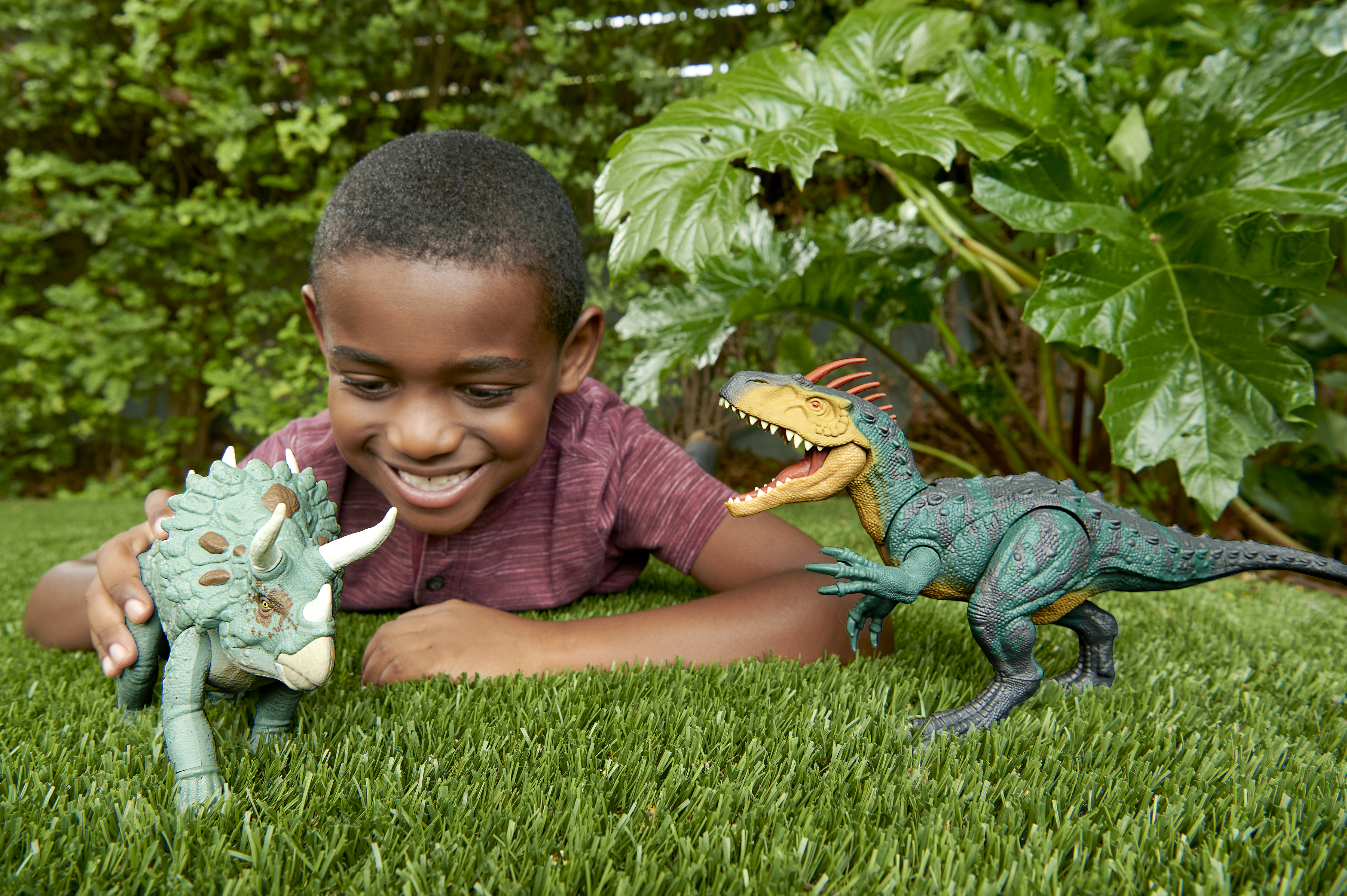 Un niño sonríe, jugando con juguetes de dinosaurios sobre hierba verde. Vegetación de fondo.