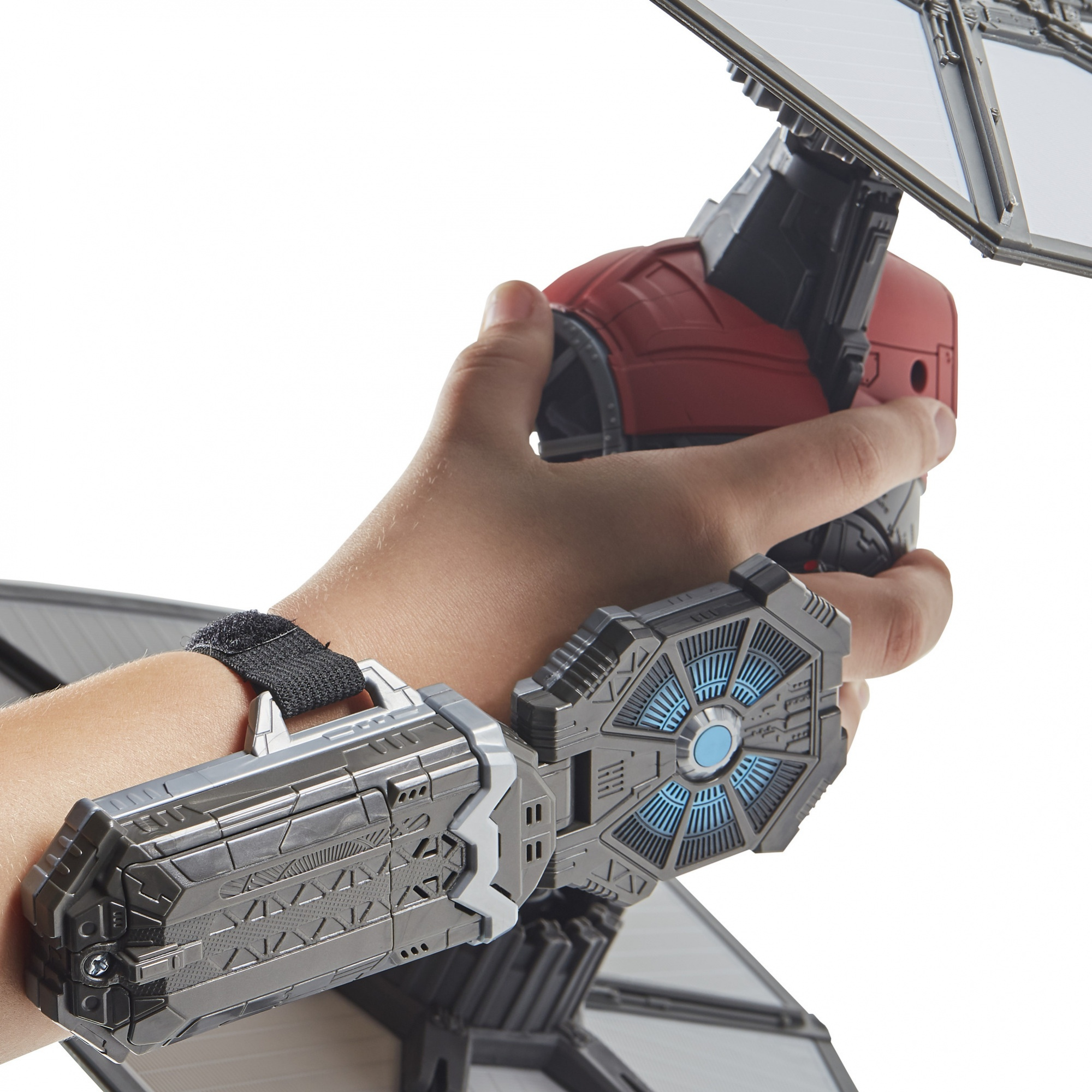 Una mano tiene un giocattolo TIE fighter. Il polso ha un dispositivo montato sul polso. Il giocattolo è grigio, nero e rosso, con uno sfondo bianco.