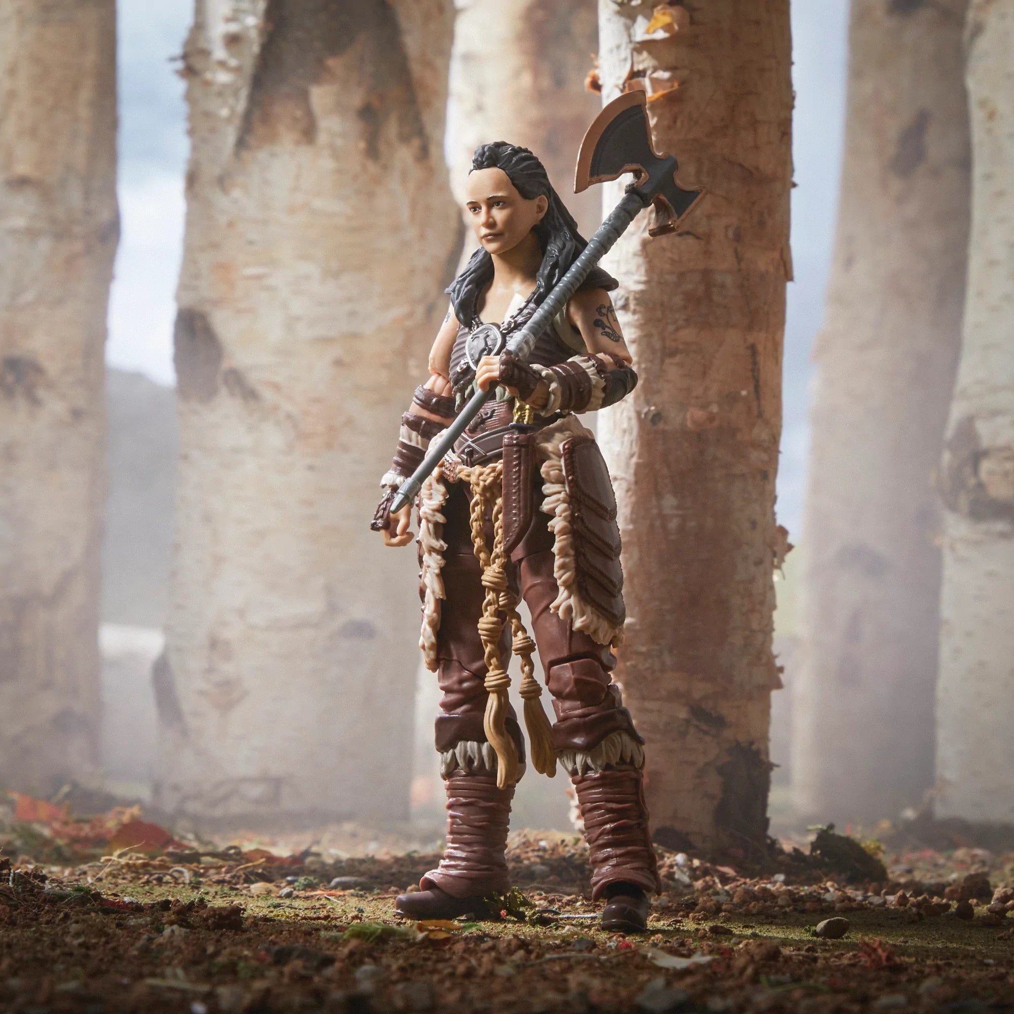 Una statuetta di guerriera femminile si trova in una foresta, con un'ascia. Indossa un'armatura marrone e si trova tra betulle.