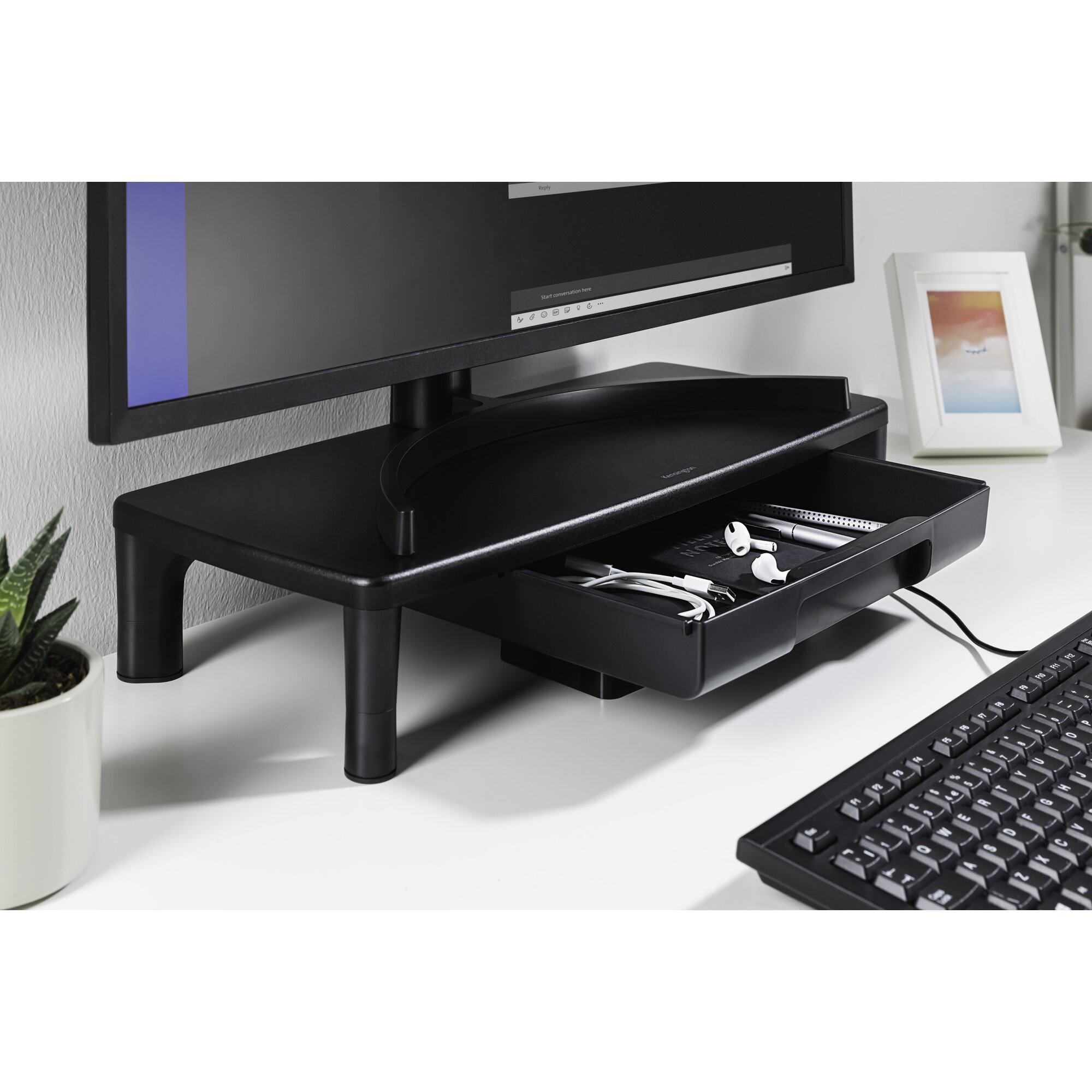 Supporto per monitor nero con cassetto su una scrivania. Tastiera, pianta, cornice e monitor sono visibili.