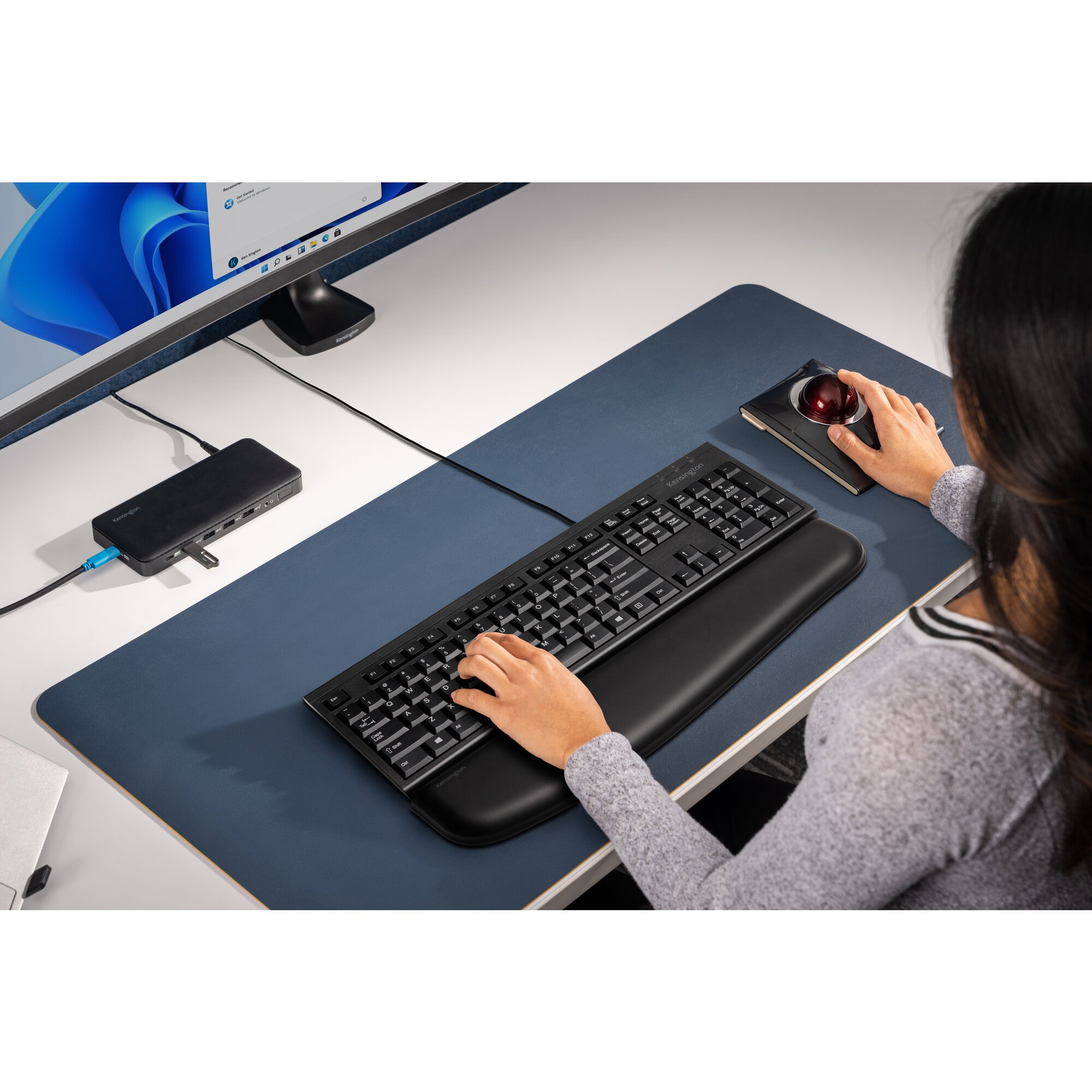 Eine Person benutzt eine Tastatur und einen Trackball. Auf dem Schreibtisch stehen ein Monitor und eine Dockingstation.