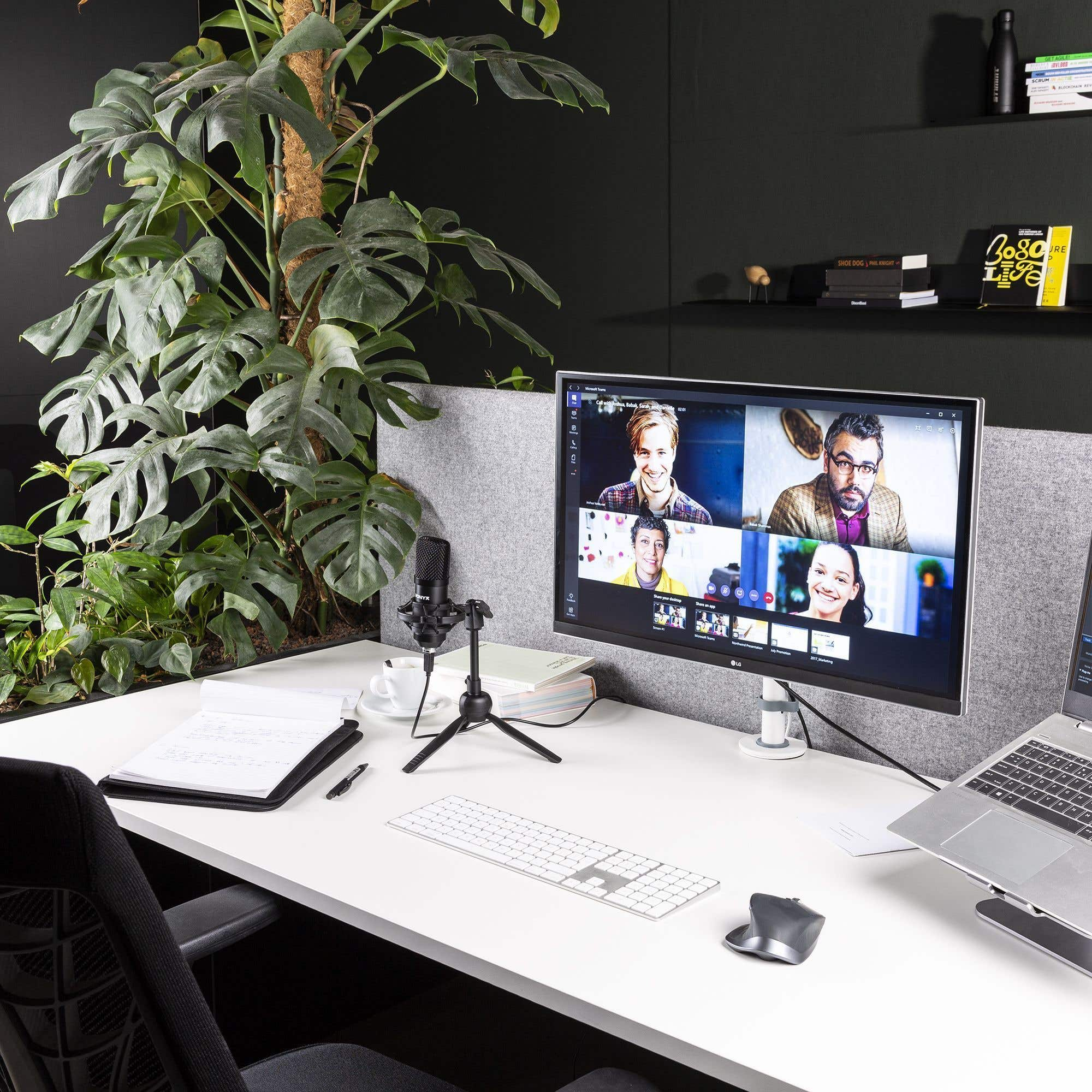Kantooropstelling met videogesprek op een monitor, microfoon, laptop, toetsenbord en grote plant.