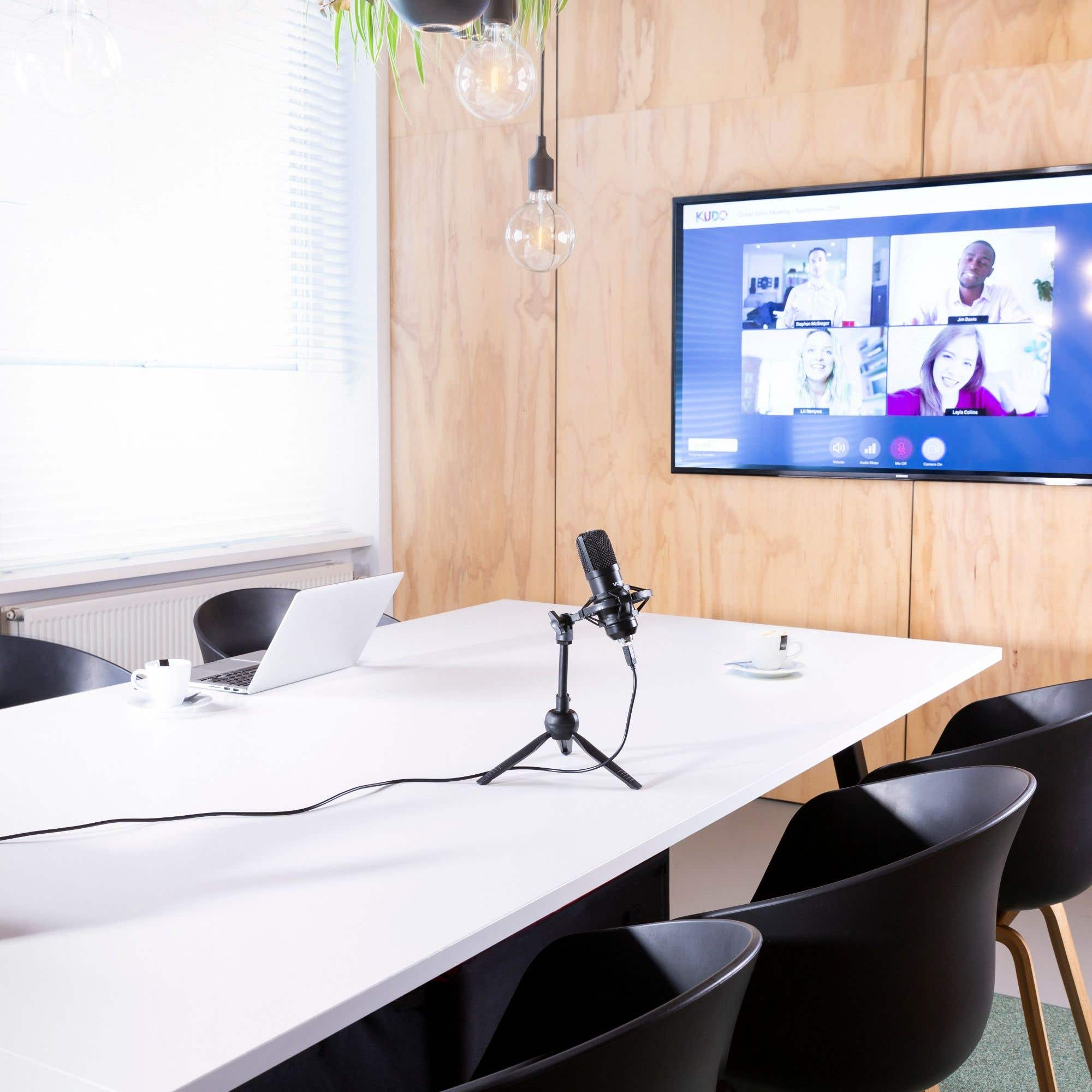 Moderne vergaderruimte. Microfoon op witte tafel, scherm met videogesprek. Zwarte stoelen eromheen.