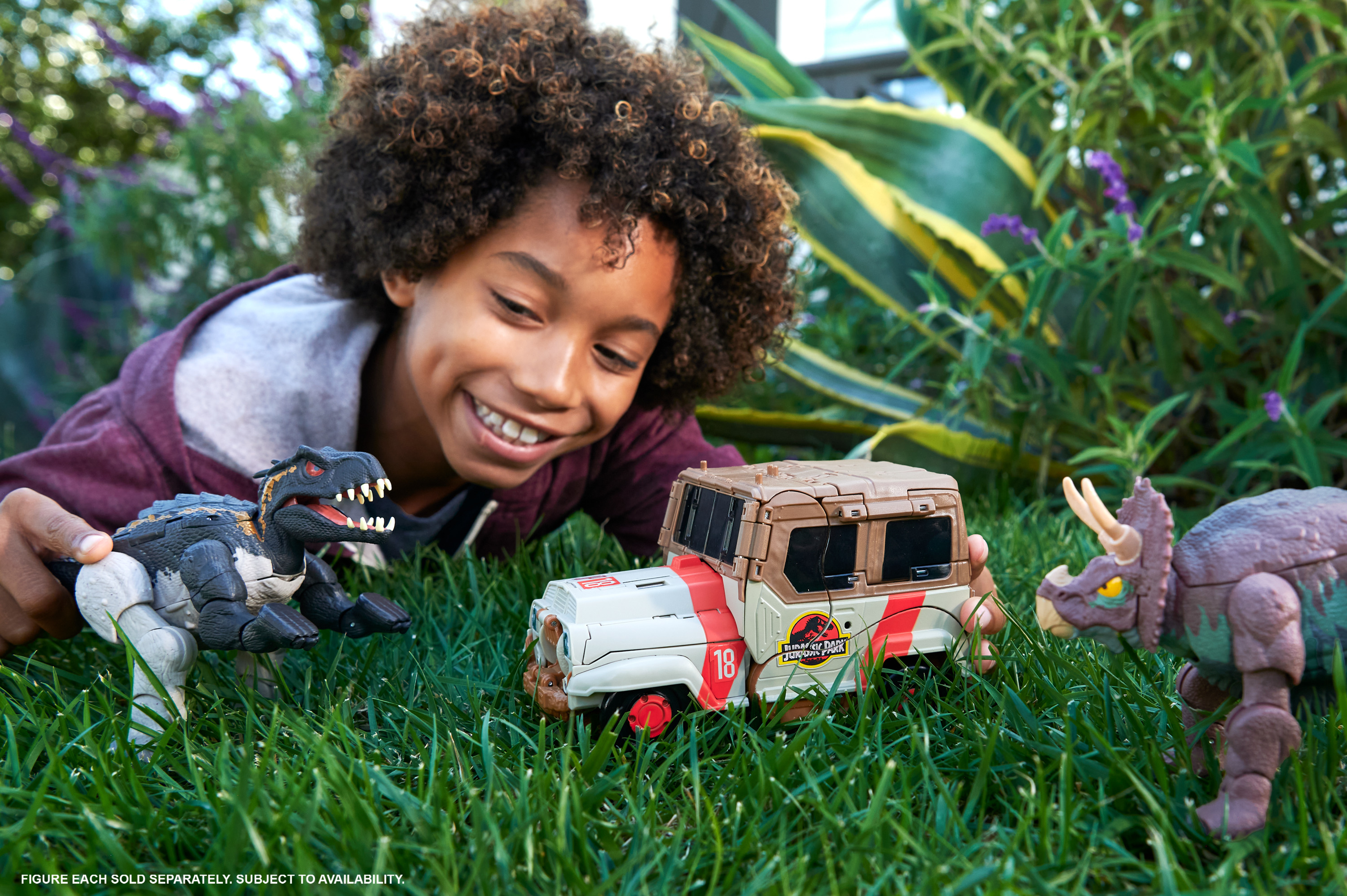 Ein lächelnder Junge spielt mit Spielzeugdinosauriern und einem Jurassic Park Jeep auf Gras. Der Hintergrund hat grüne Pflanzen.