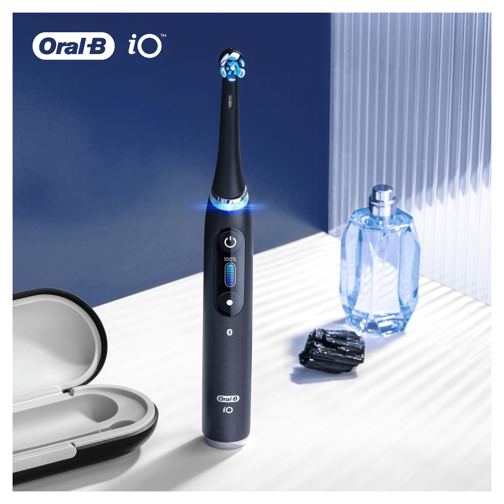 Schwarze Oral-B iO Zahnbürste mit blauem Licht, Parfümflasche und einem schwarzen Stein auf weißer Oberfläche.