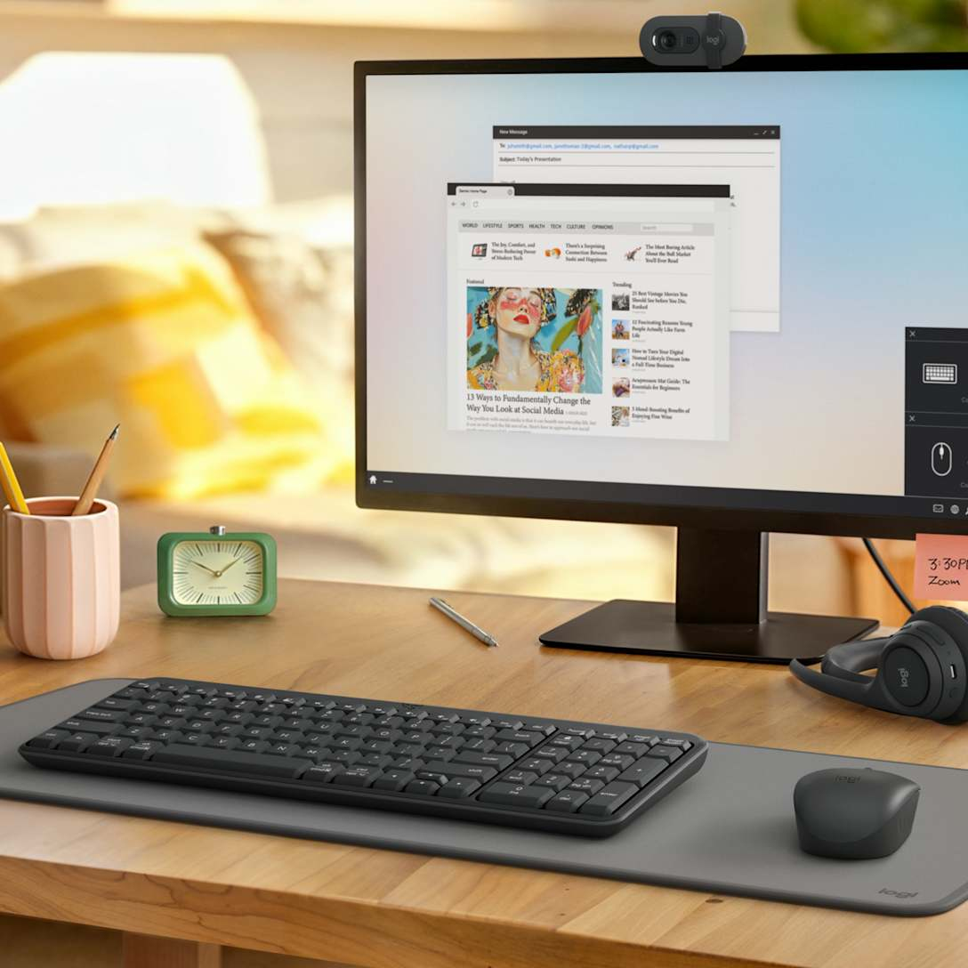 Configurazione scrivania con monitor, tastiera, mouse, webcam, orologio, portapenne e cuffie.
