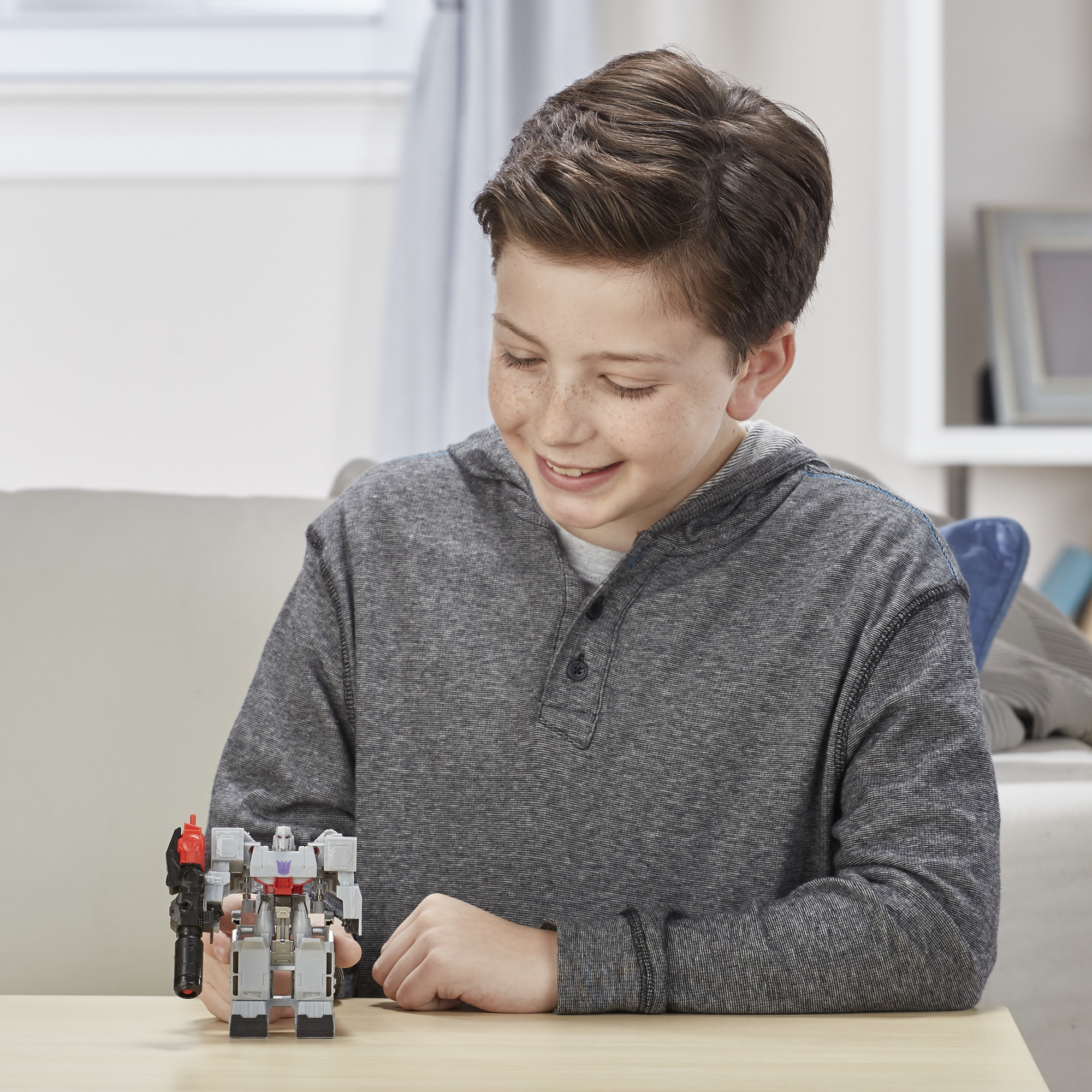 Ein Junge lächelt einen grau-schwarzen Roboterspielzeug an. Er trägt einen grauen Kapuzenpullover. Das Spielzeug steht auf einem Holztisch.