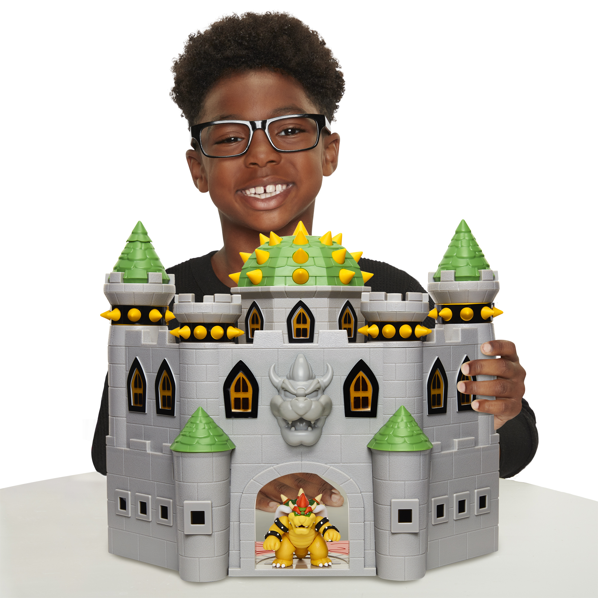 Ein Junge mit Brille hält eine Spielzeugburg. Die Burg ist grau, mit grünen und gelben Akzenten. Bowser befindet sich in der Burg.
